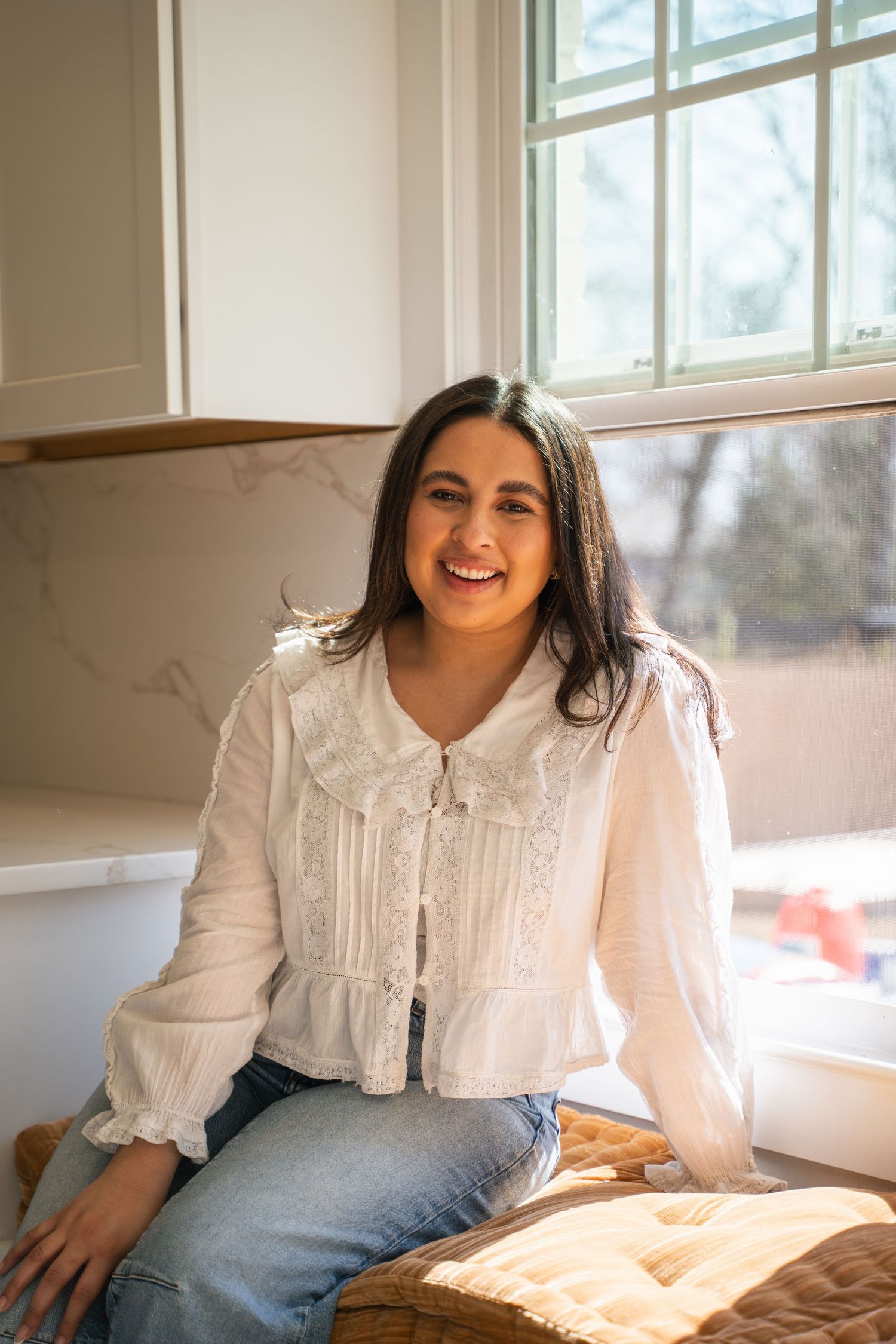 Smiling young woman in white lace blouse sitting by a bright window in a cozy indoor setting.