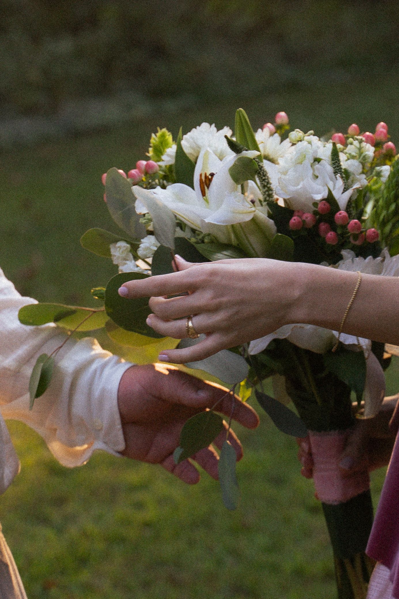 Close-up of bride and groom's hands touching a wedding bouquet of white lilies and pink buds.
