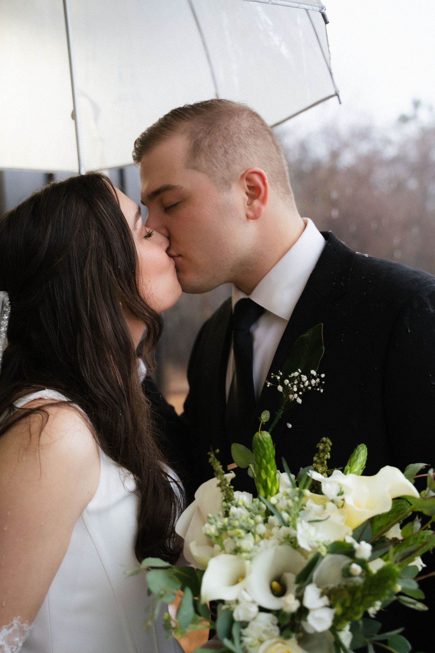 Bride and groom sharing romantic kiss under clear umbrella on rainy wedding day.