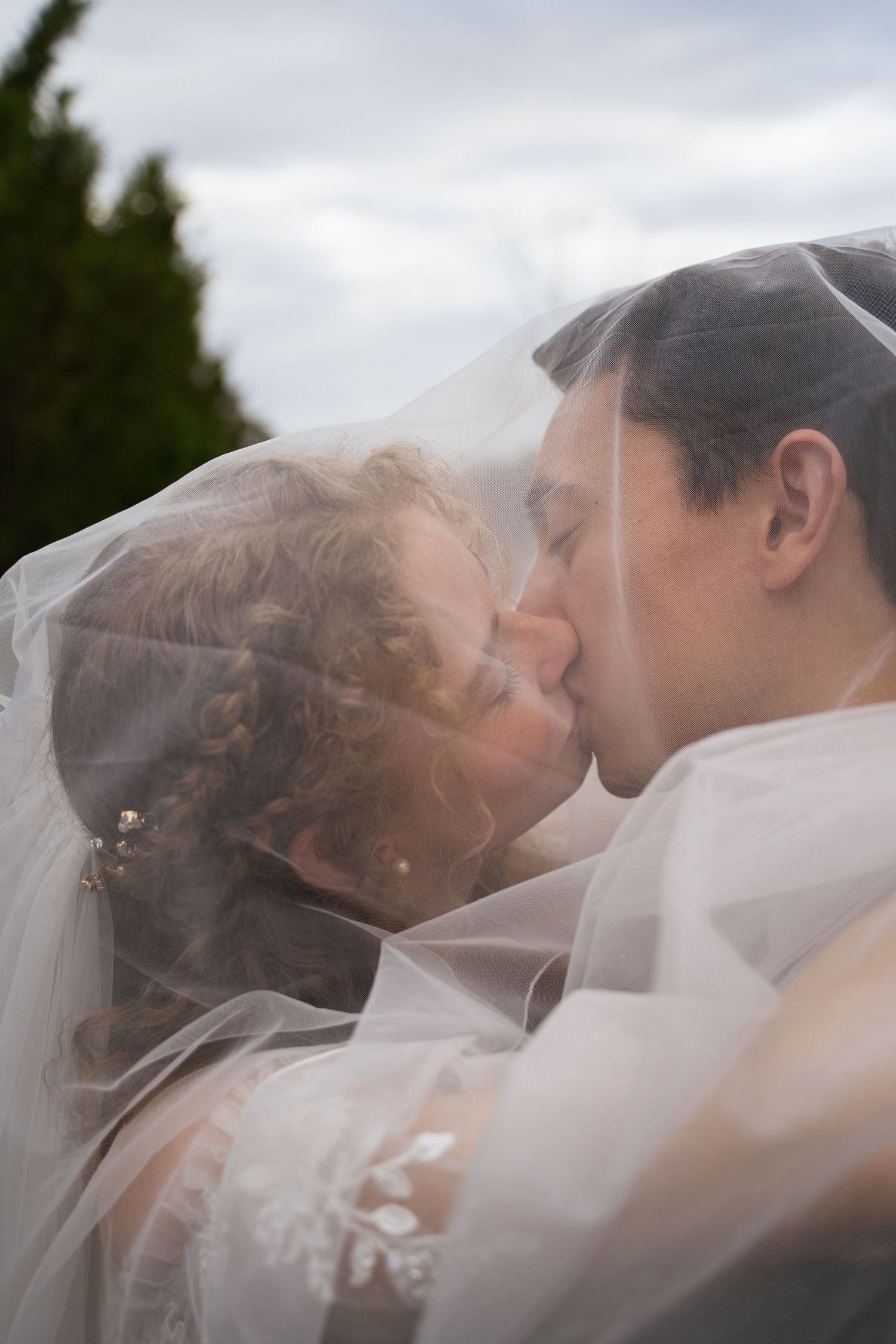 Bride and groom share romantic kiss under flowing white veil outdoors on their wedding day.