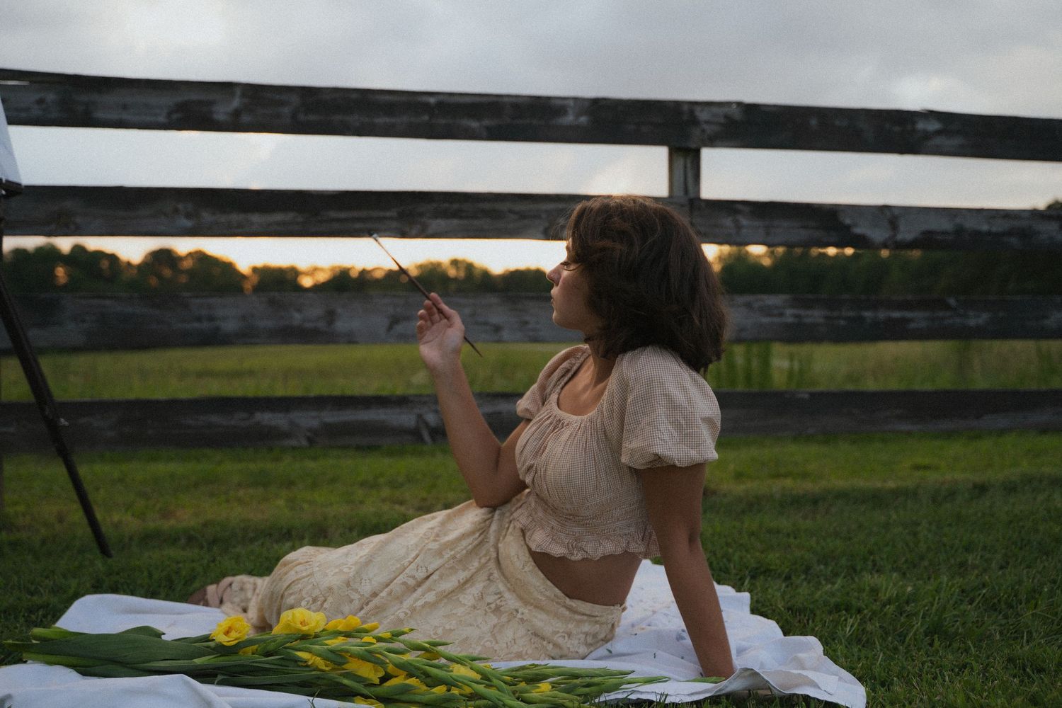 a young woman sits outside on a picnic blanket with flowers holding a paintbrush and looking away