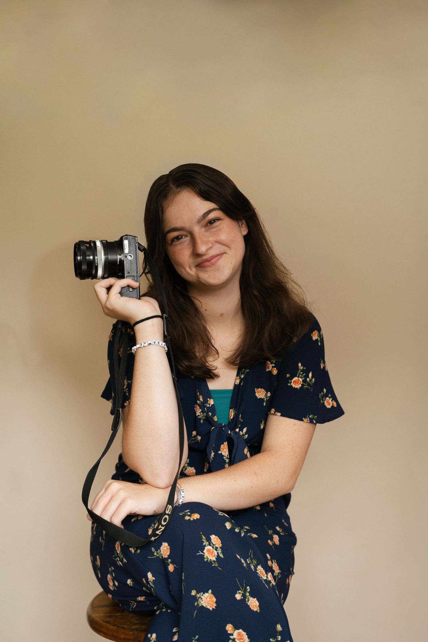 Young woman in floral dress holding a vintage film camera while sitting on a stool.