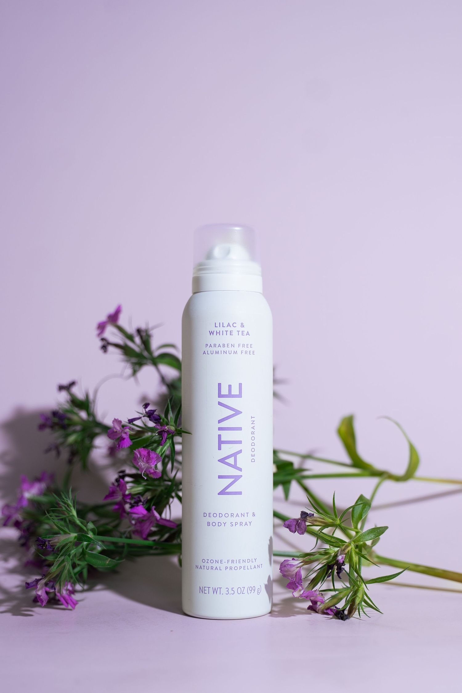 Native Lilac and White Tea deodorant body spray surrounded by purple wildflowers on lavender background.