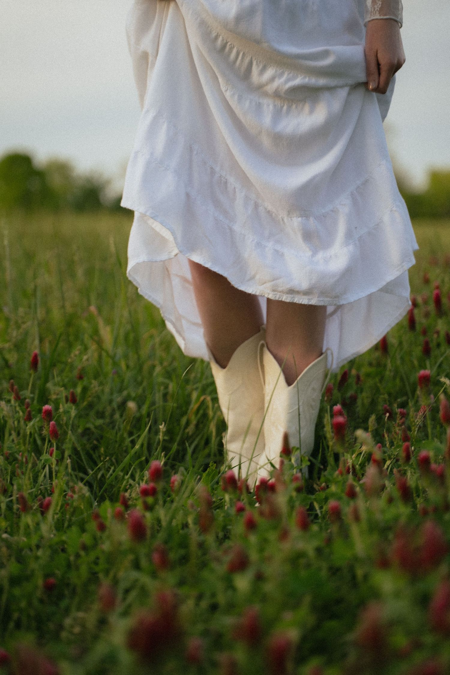 Woman in white dress and cowboy boots walking through a field of red clover flowers.
