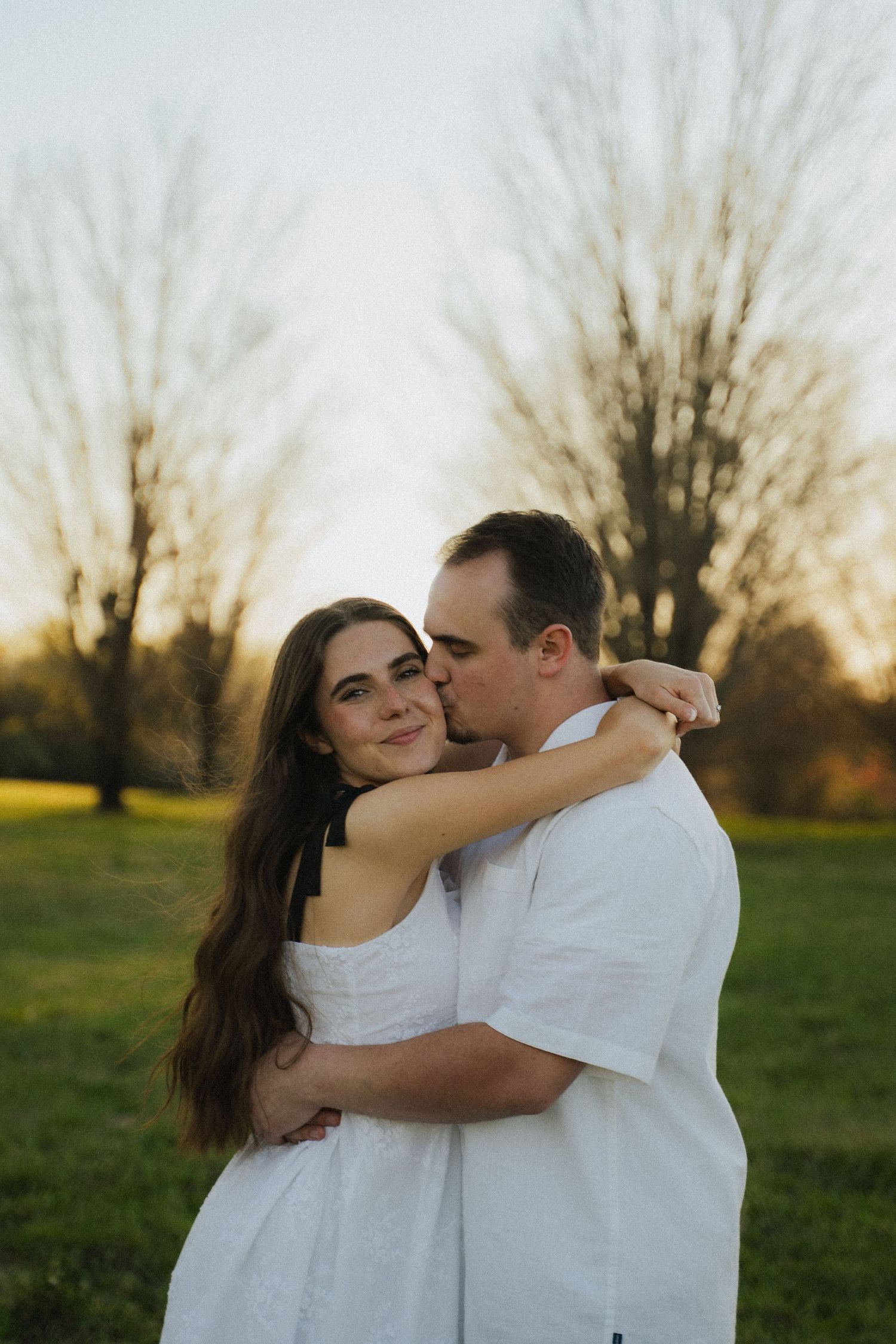 Man kisses woman on cheek as she smiles in a golden sunset park engagement photo.