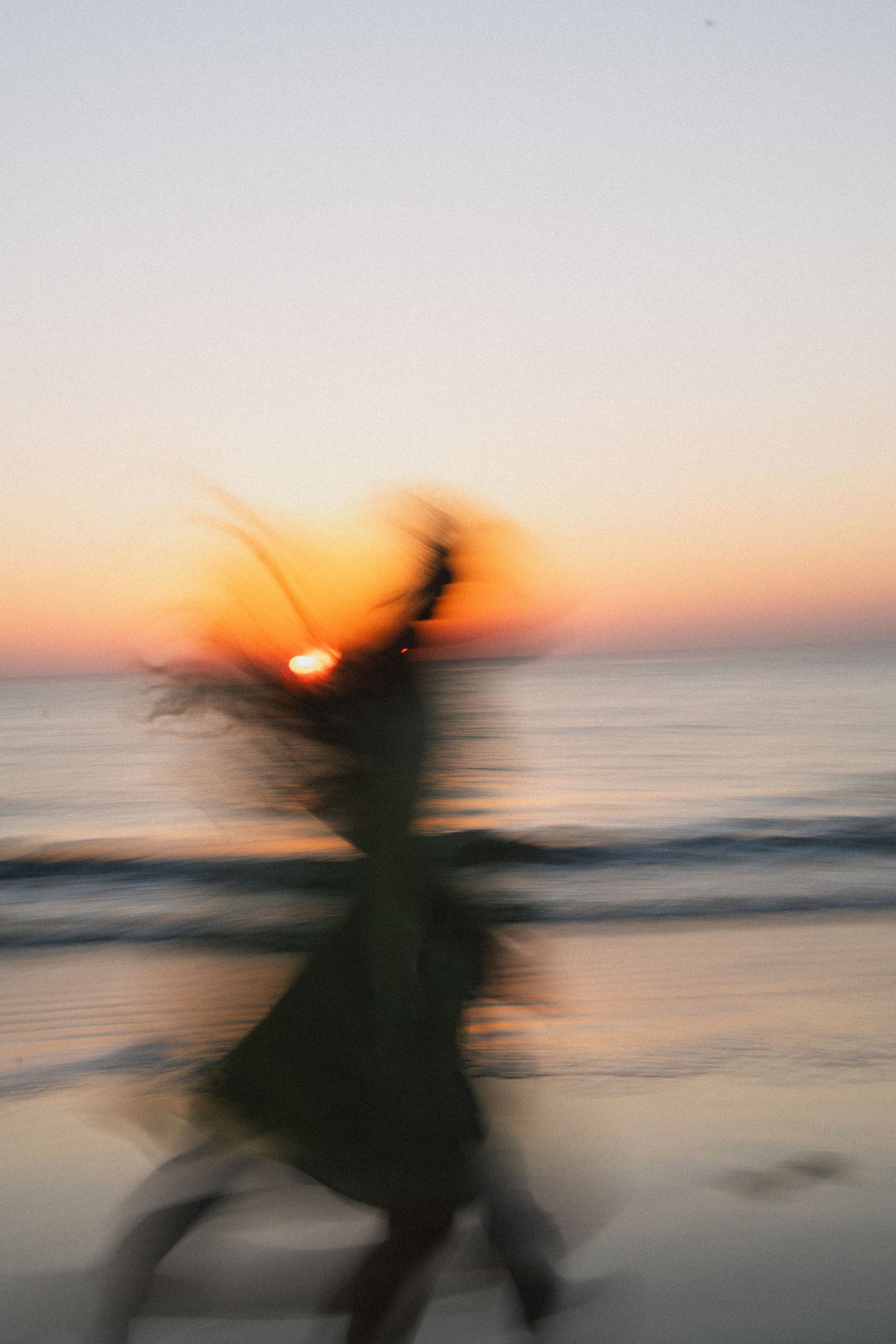 Blurred motion photo of a woman dancing on a beach at sunset with golden orange sky.