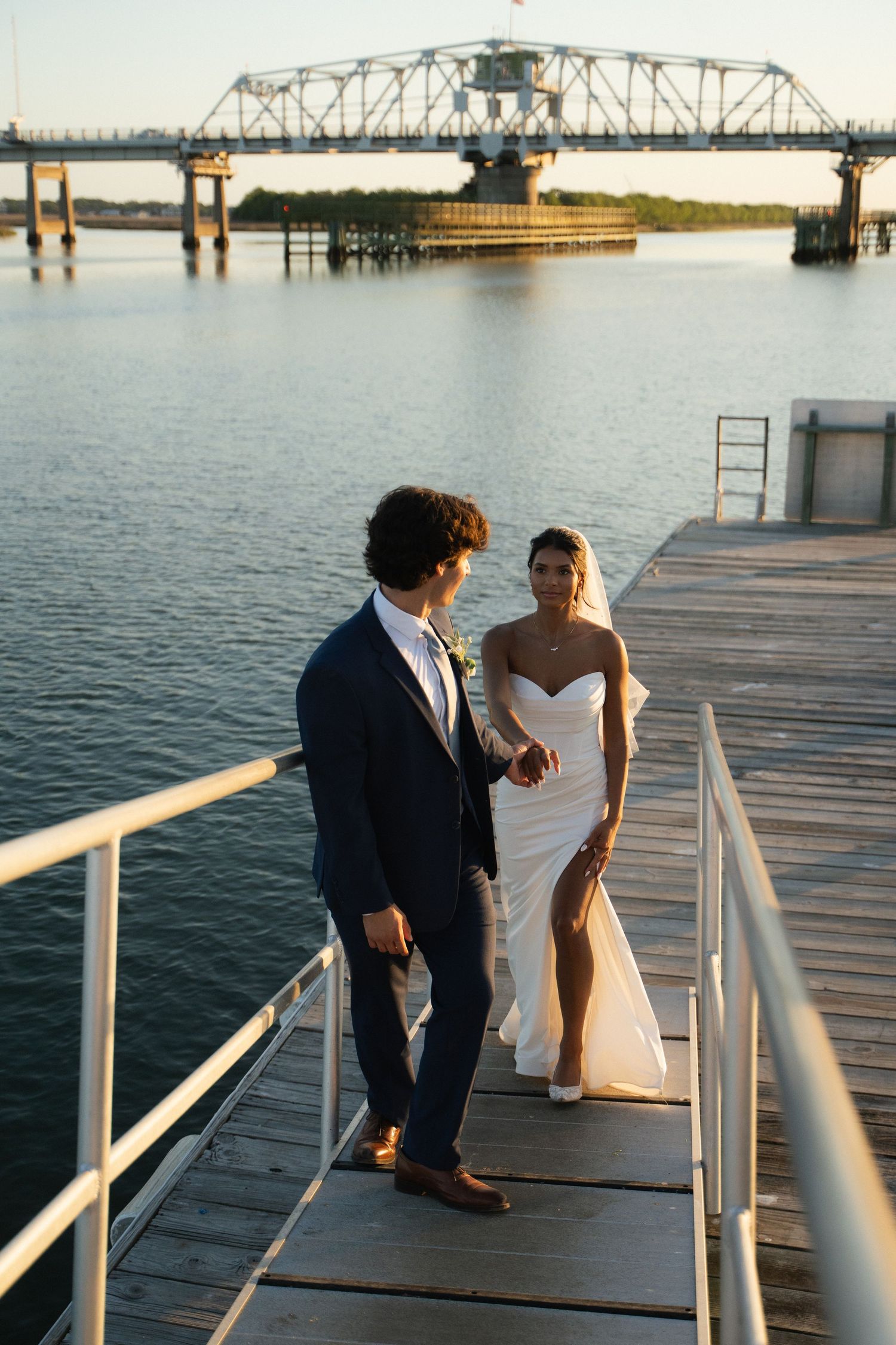 Bride and groom walk hand in hand on waterfront dock at golden hour with bridge backdrop.
