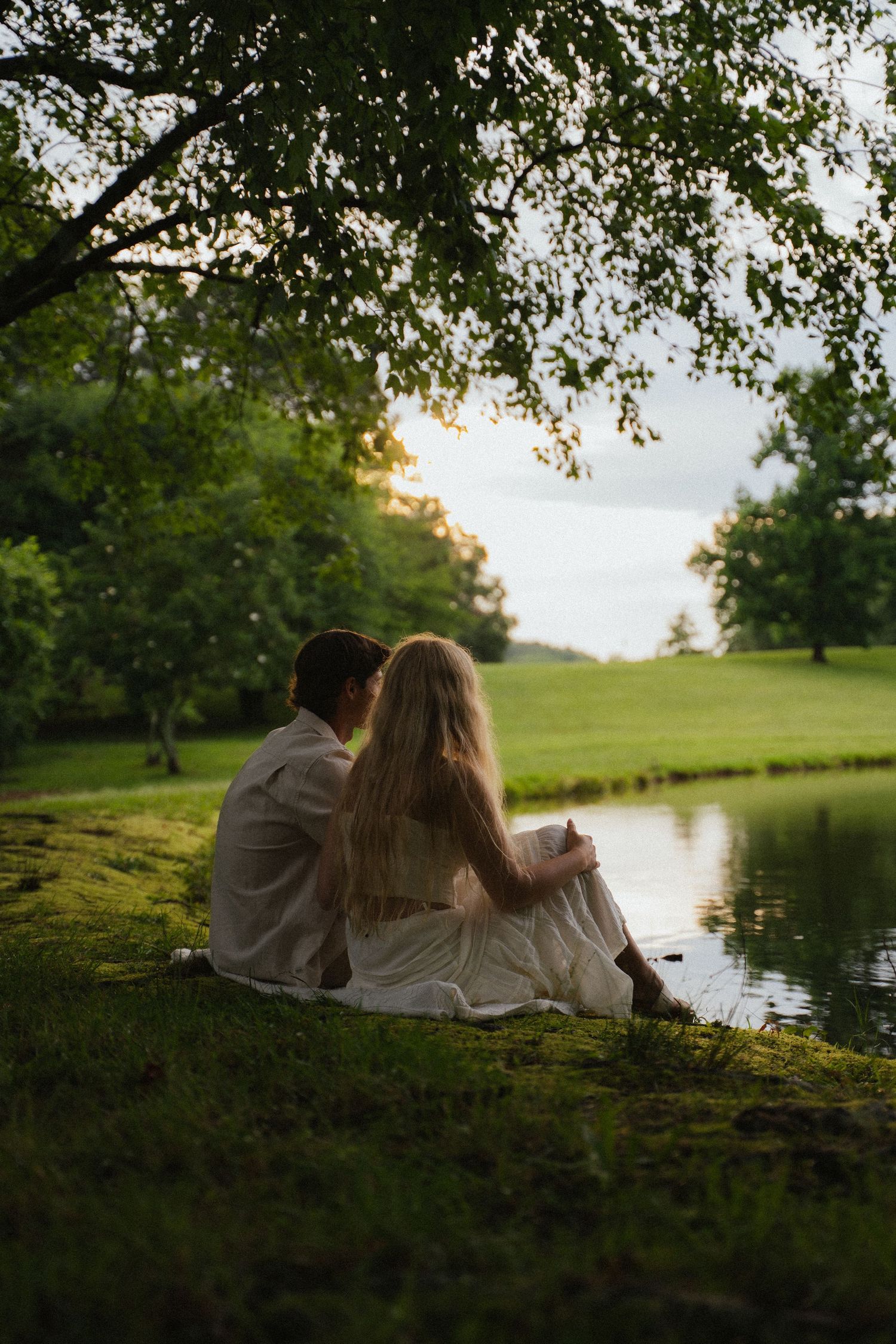 Couple sits together on grassy riverbank under tree canopy at golden hour sunset.