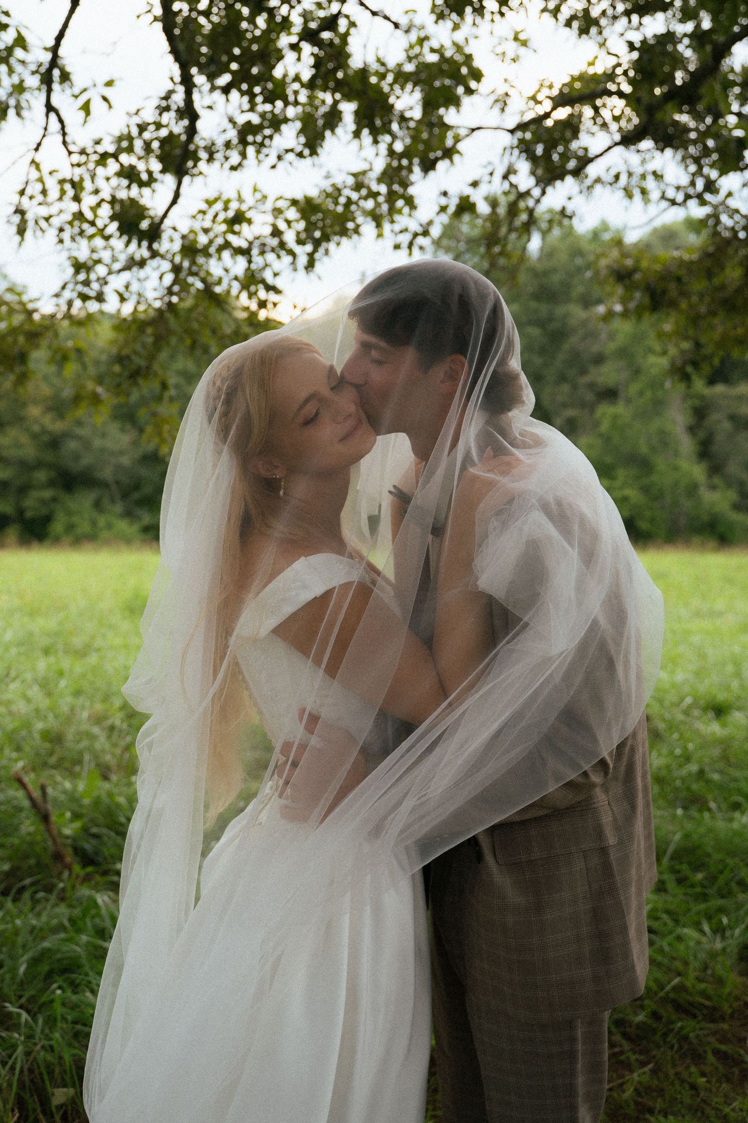 Bride and groom sharing romantic kiss under flowing veil in lush green outdoor setting.