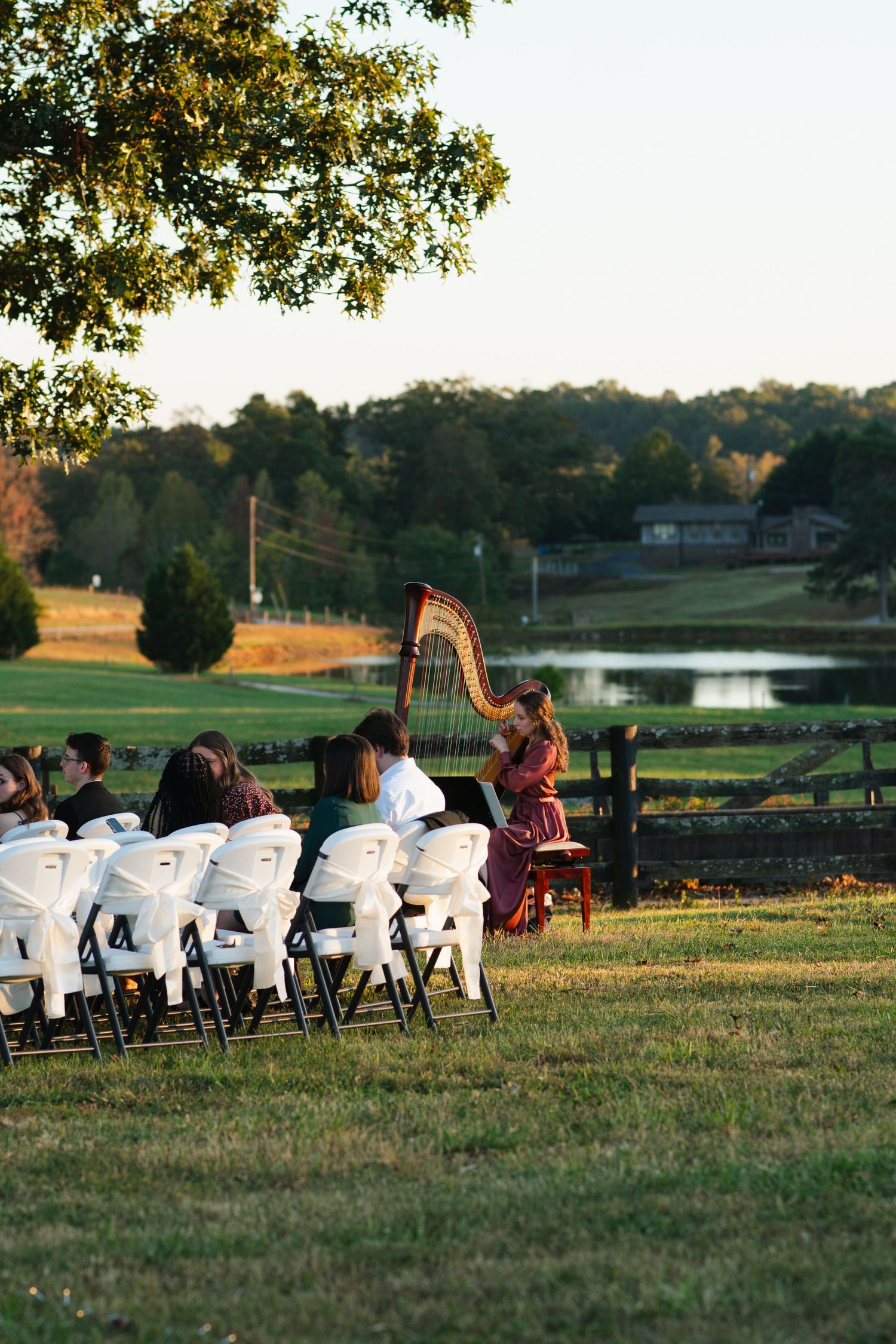 Harpist in red dress performs for seated wedding guests at scenic outdoor ceremony near pond.