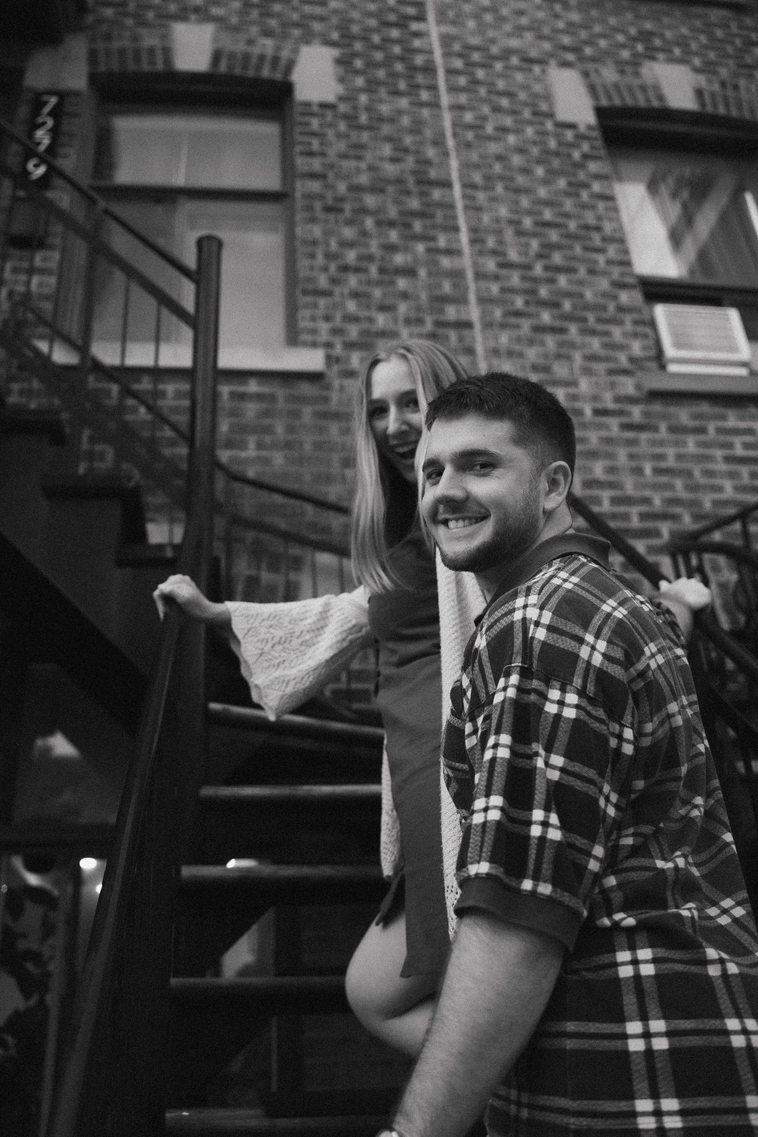 Black and white photo of a smiling couple on urban apartment fire escape stairs at night.