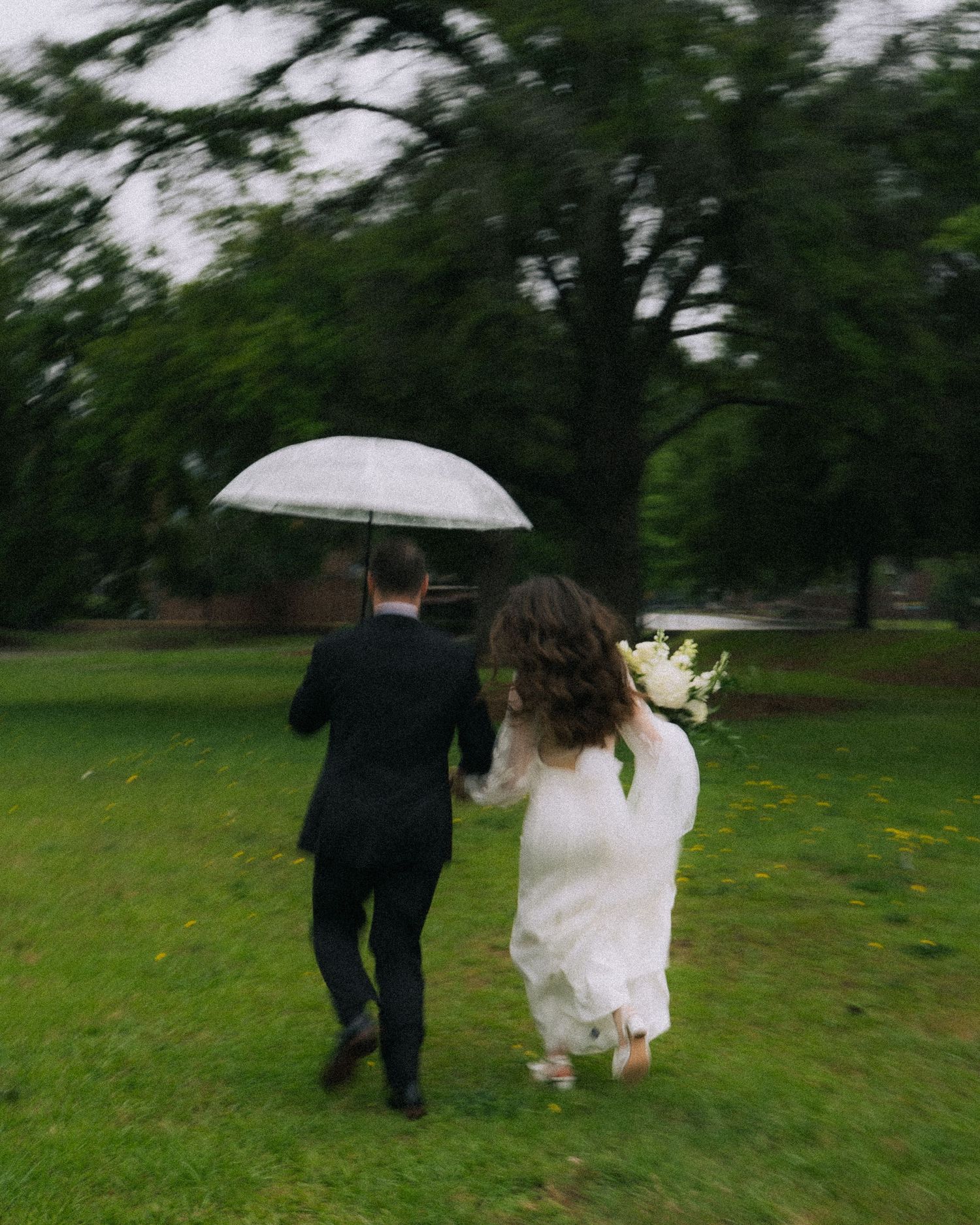 Bride and groom running across a green lawn in the rain, sharing a clear umbrella with white bouquet.