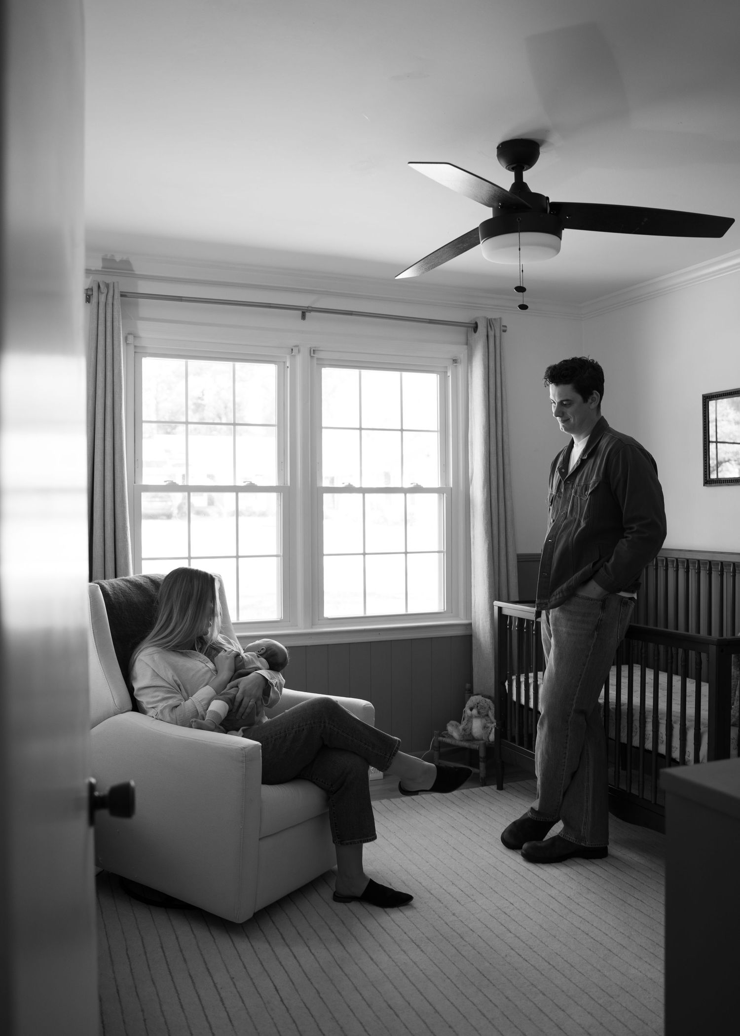 Black and white photo of new parents with newborn baby in a nursery with crib and ceiling fan.