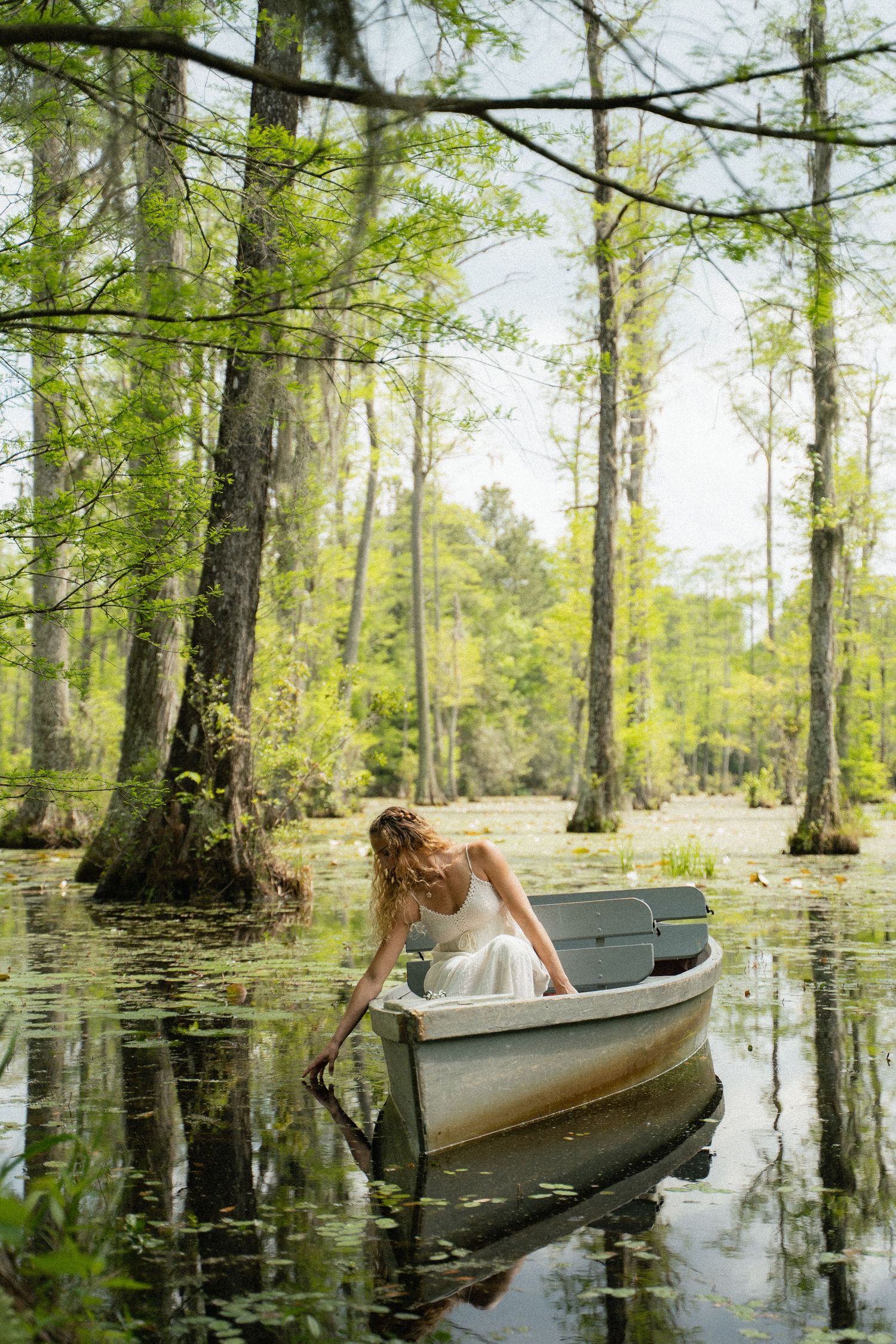 Woman in white dress reaching into water from a boat in a serene cypress swamp.