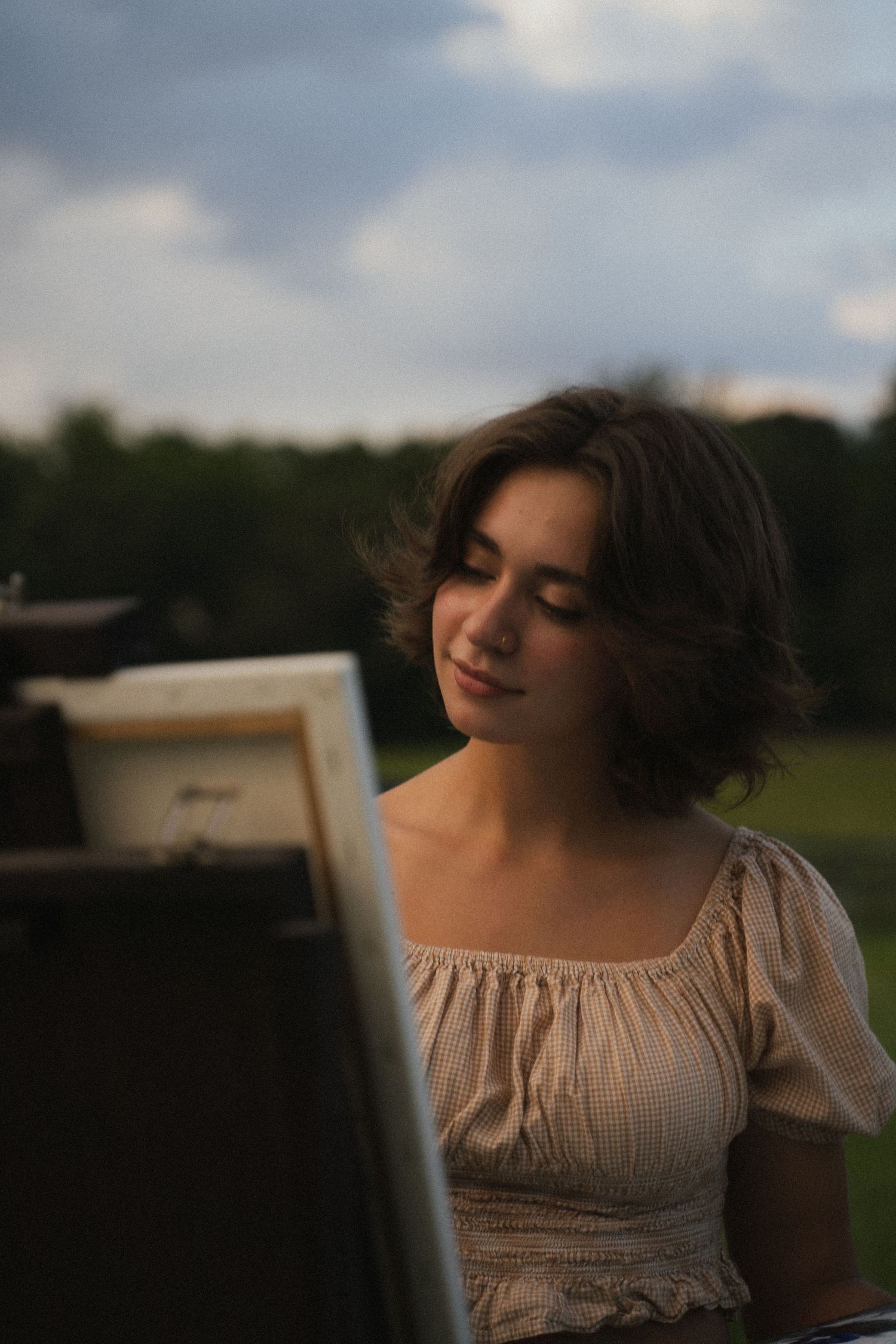 A young brunette woman in a beige top smiles softly while looking at a canvas on an easel outdoors.