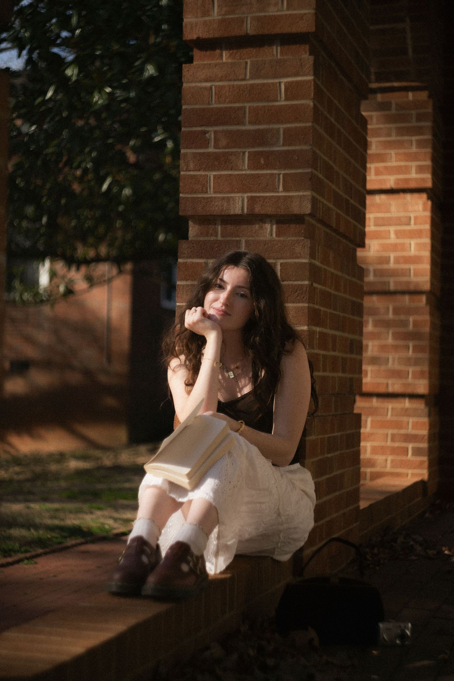 Young woman with dark curly hair sitting by brick column reading a book outdoors.