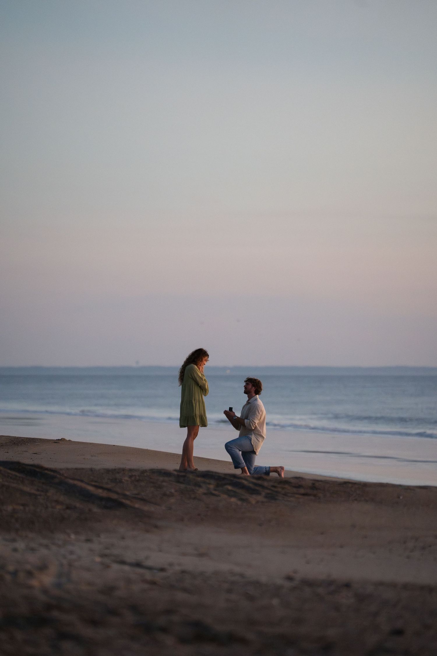 Man proposing on one knee to woman in green dress on a beach at dusk.