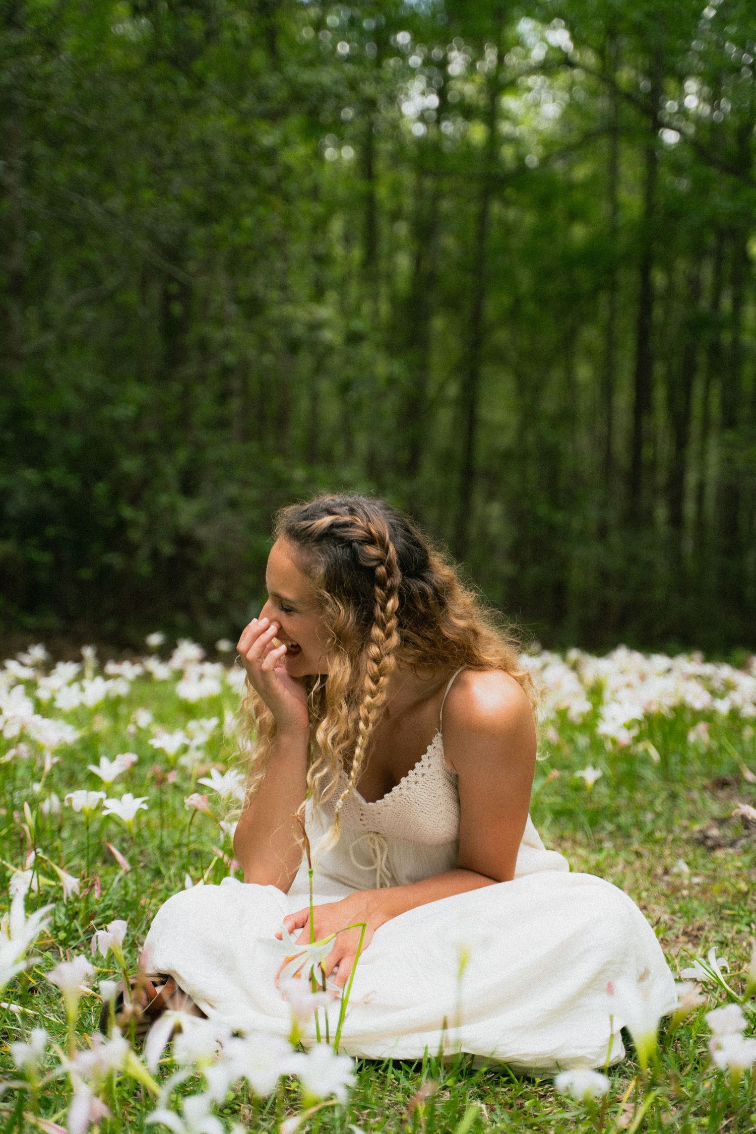 Young woman in white dress laughing among white wildflowers in a lush green forest meadow.