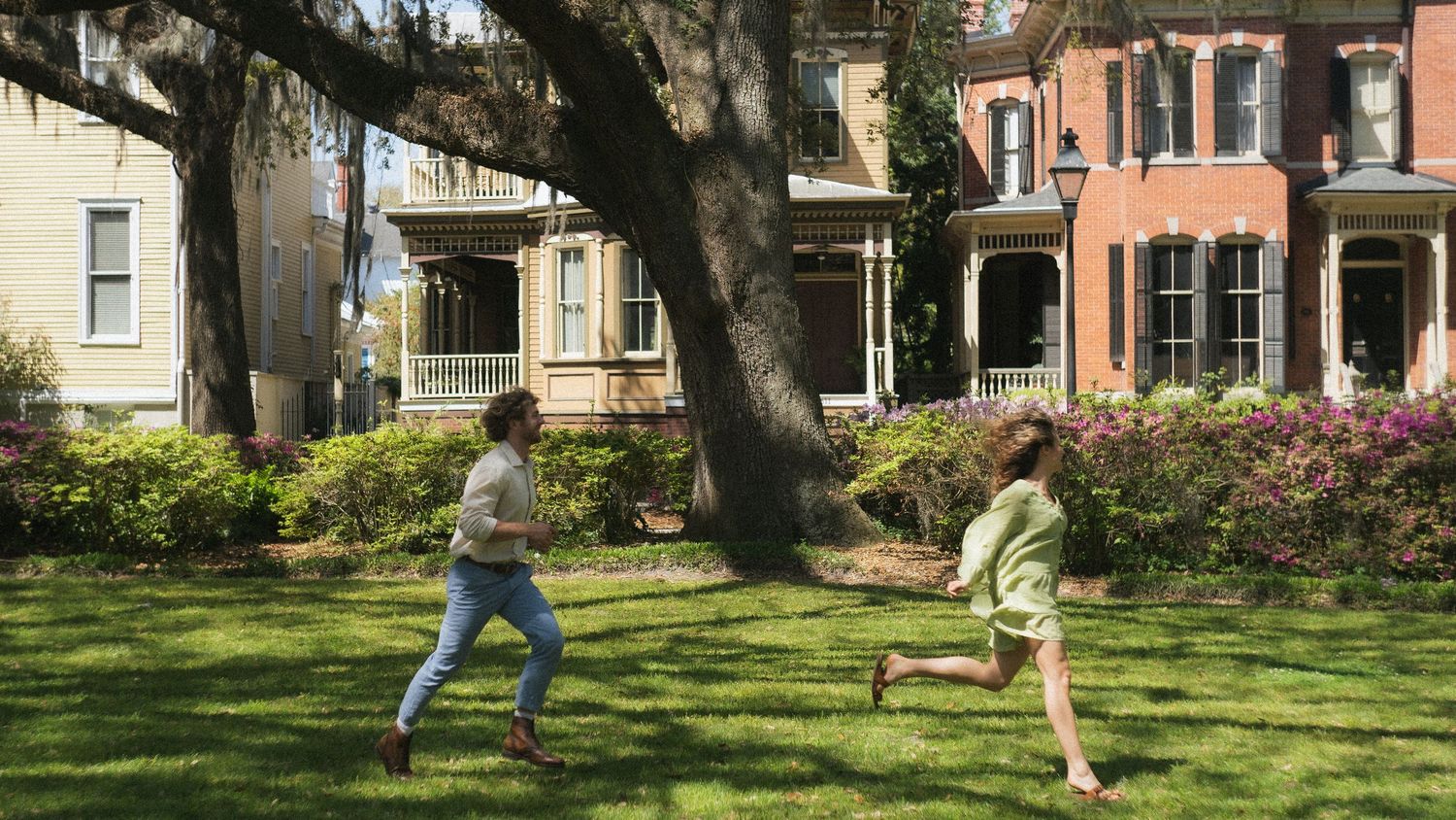 A couple playing and running in a sunny green yard in front of colorful historic townhouses.