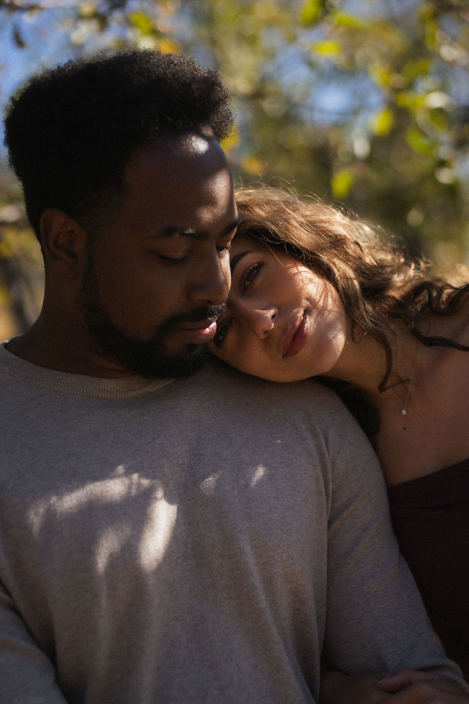 Interracial couple embracing under dappled sunlight with woman resting head on mans shoulder.