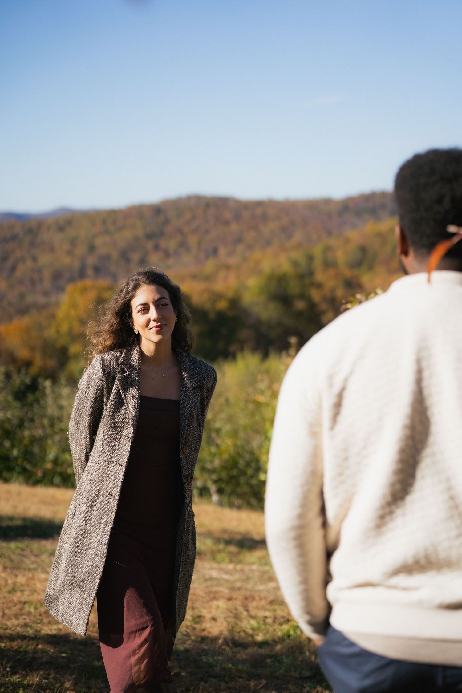 A woman in a gray coat smiles at a person in the foreground with autumn mountain scenery behind her.