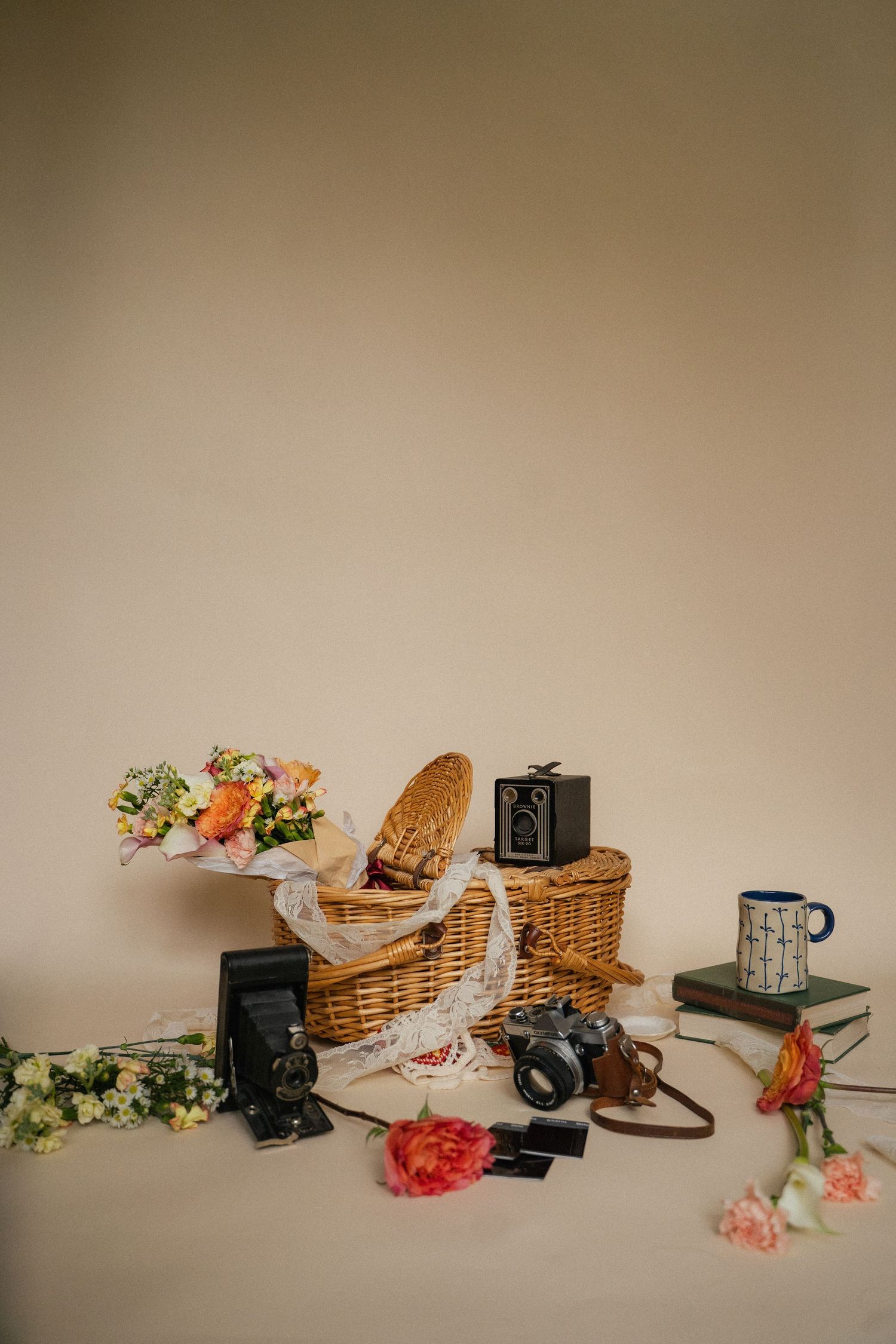 Vintage cameras, wicker picnic basket, colorful flowers, books, and mug styled on a beige background.