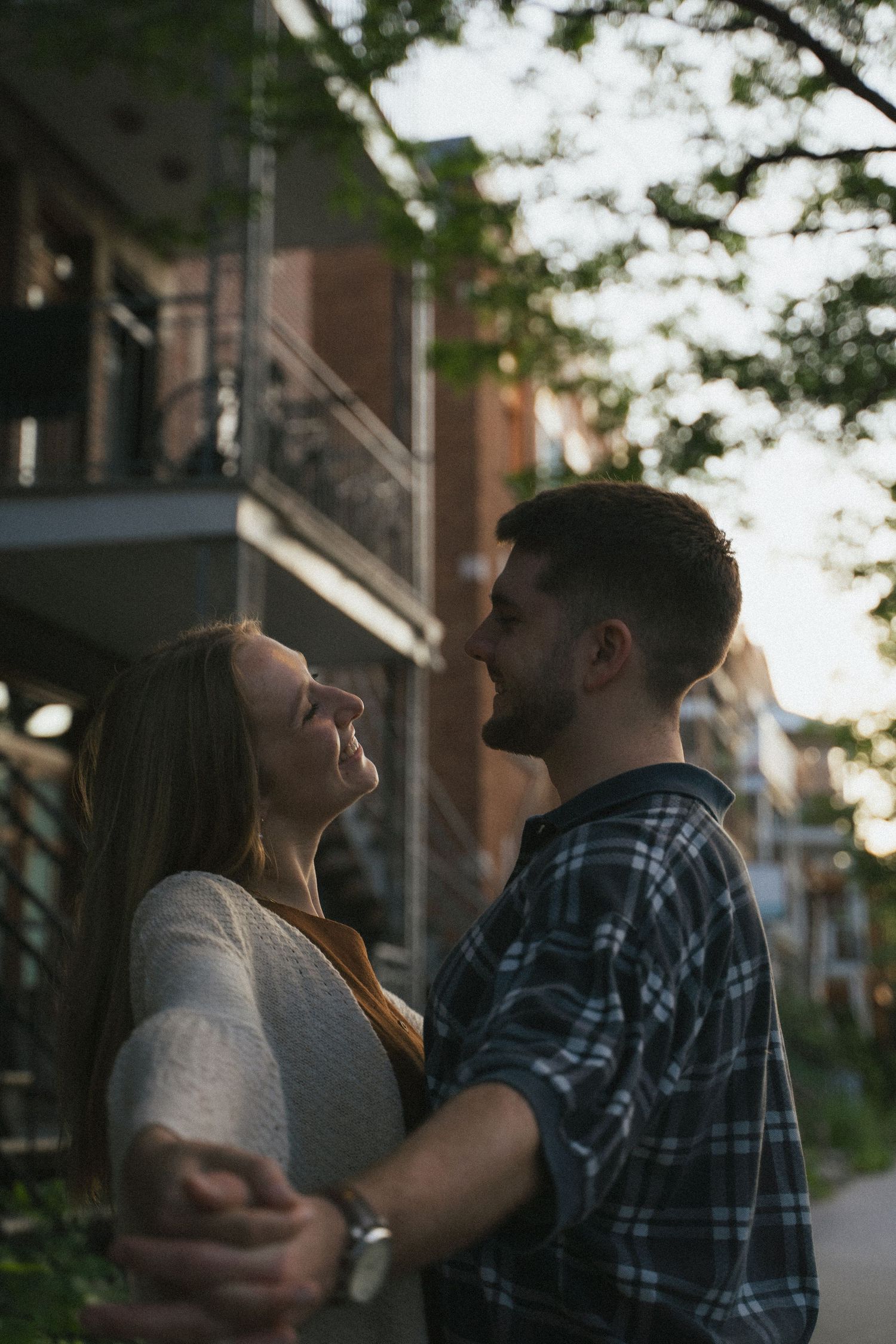 Romantic couple holds hands and gazes at each other near brick building at dusk.