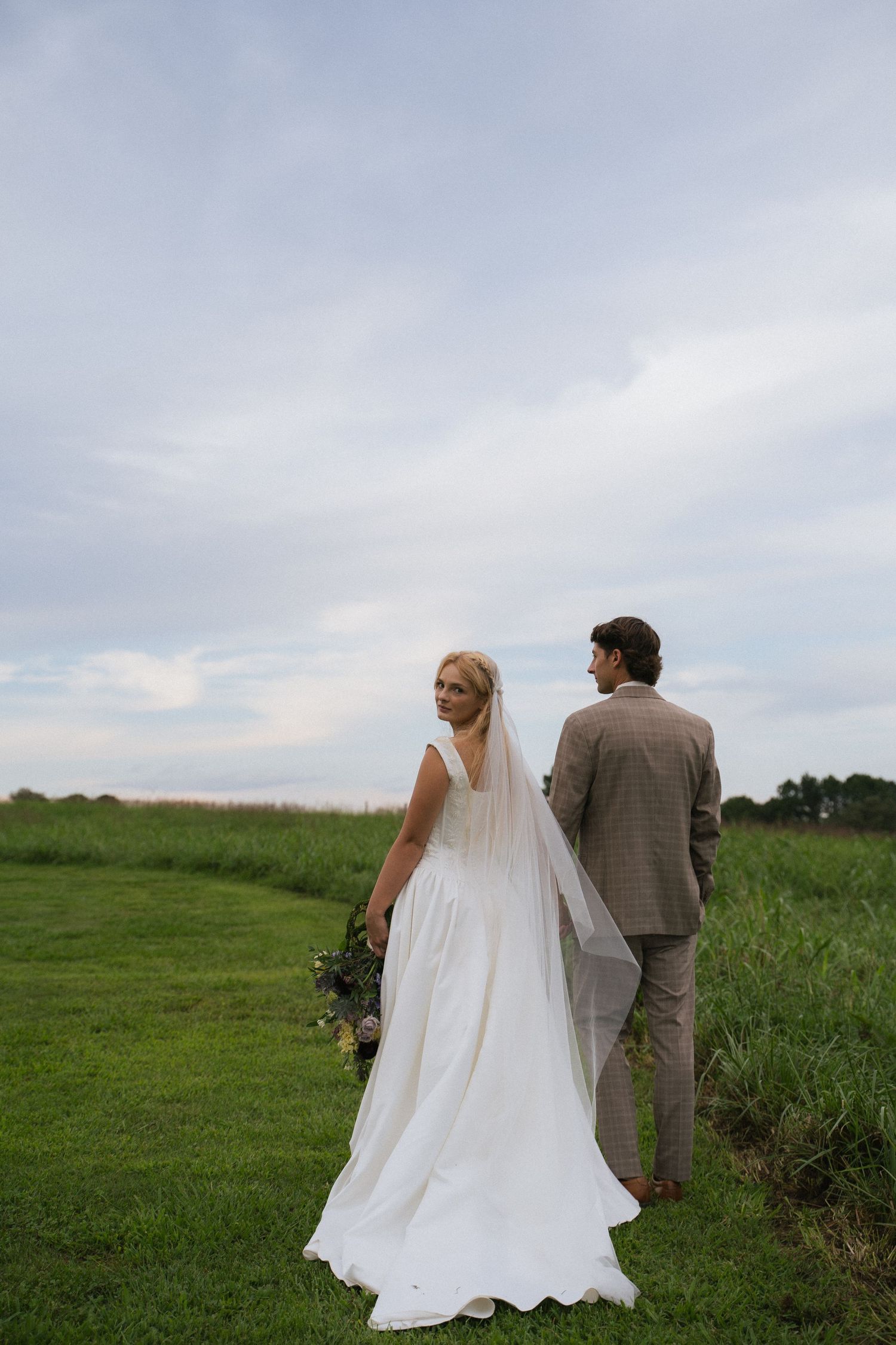 Bride in flowing white wedding gown and veil looks back while walking with groom through green countryside.