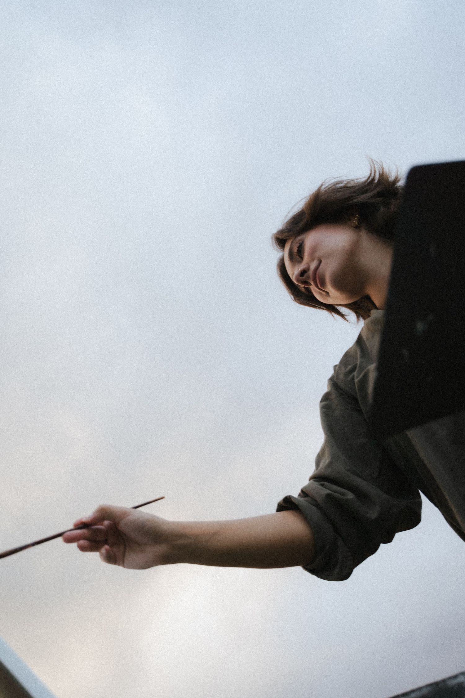 Low angle view of young woman holding a paintbrush against a cloudy sky, artistic portrait photography.