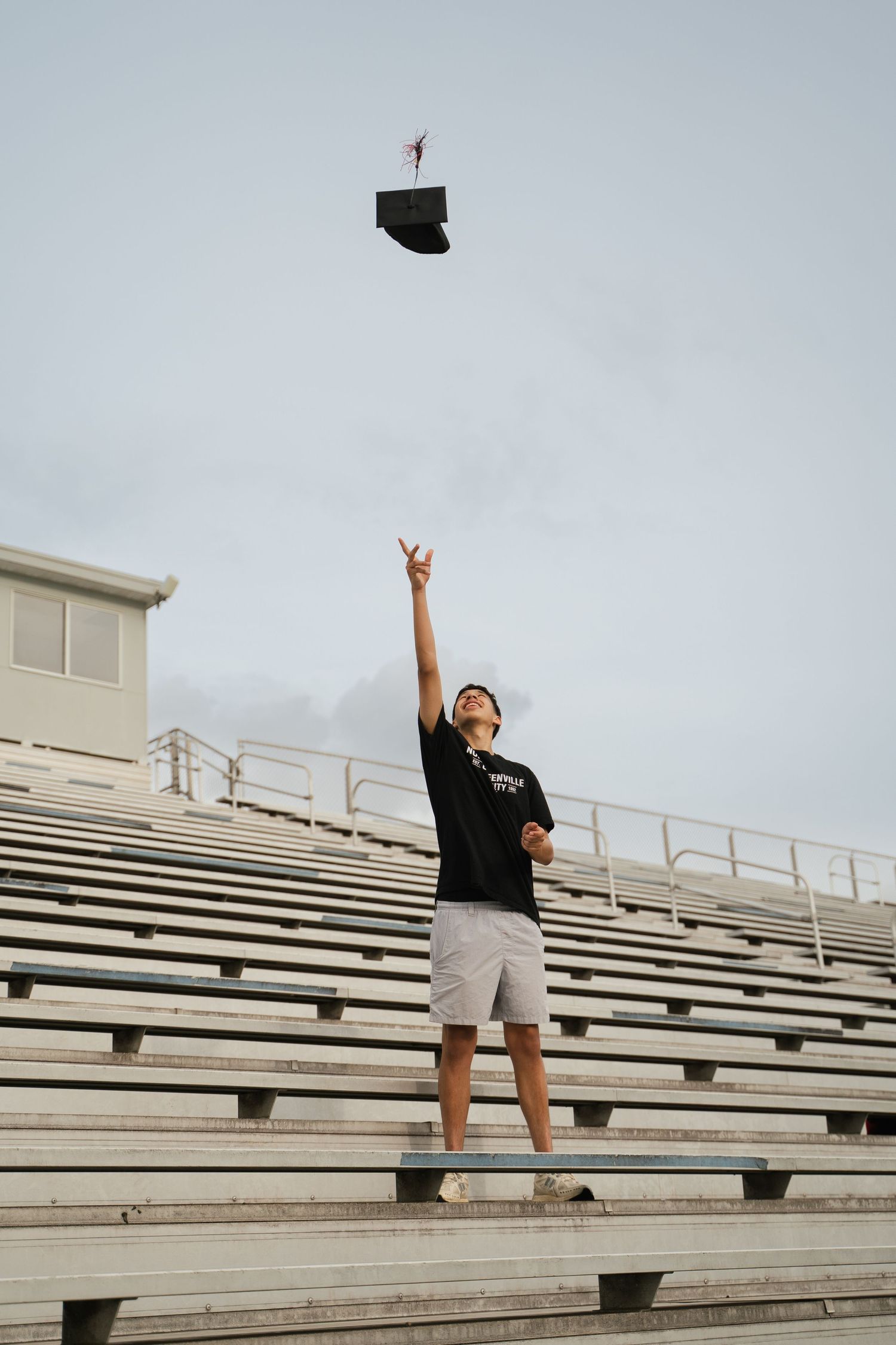 Graduate tossing cap in air while standing on bleachers against cloudy sky.