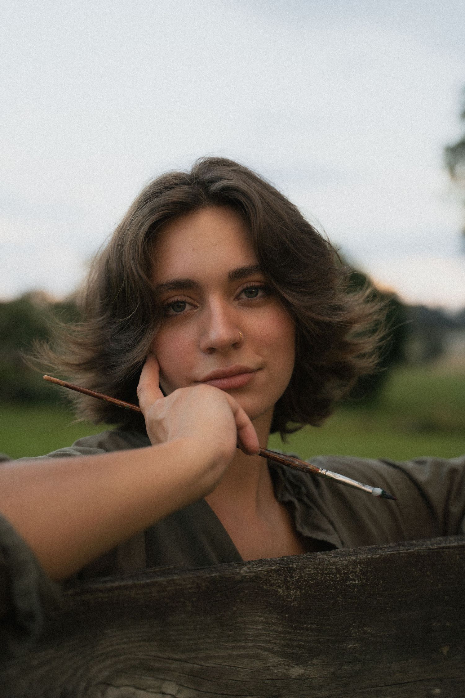 Young person rests chin on hand holding paintbrush while leaning on wooden fence outdoors.