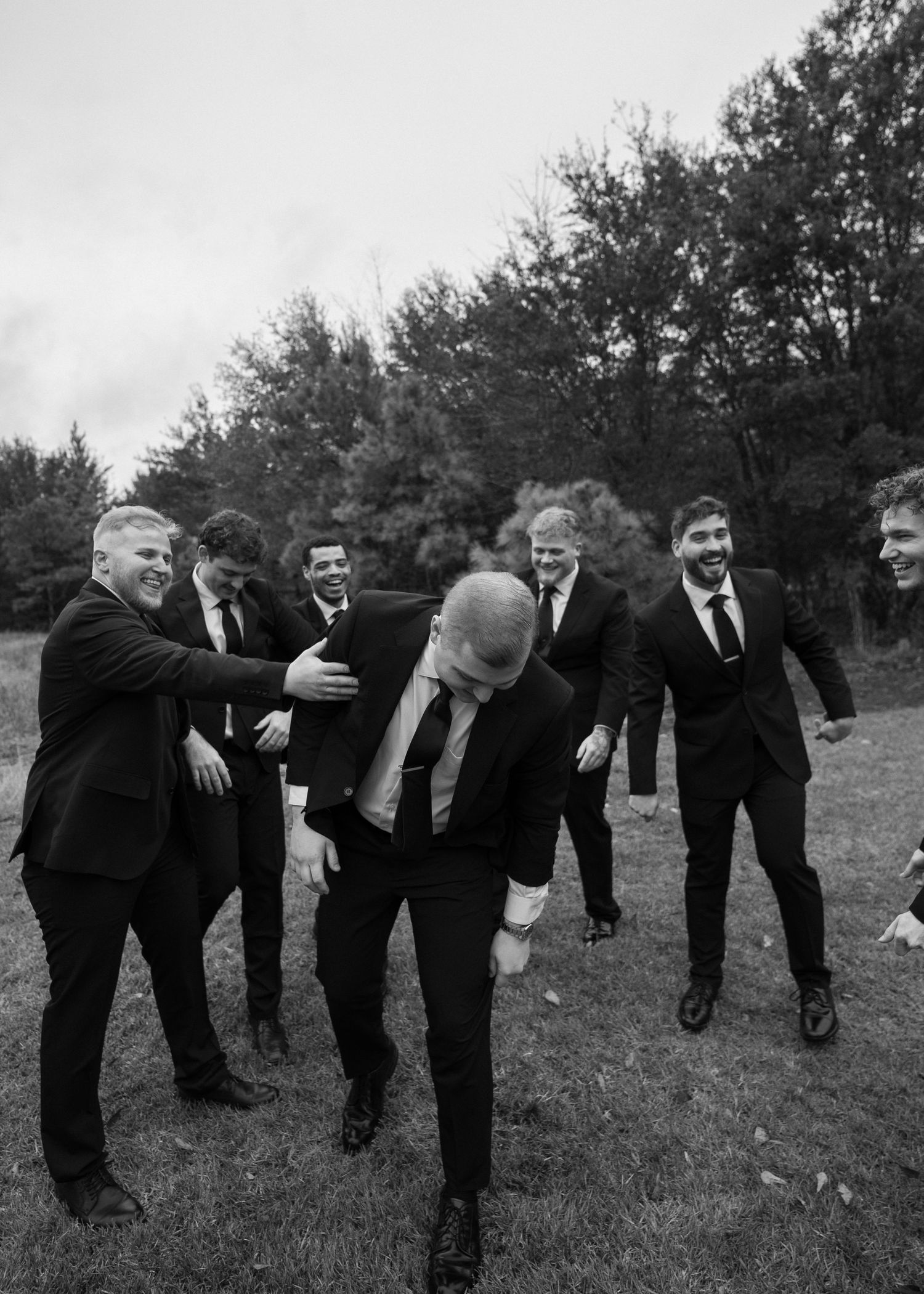 Groom laughs with groomsmen in black suits during fun candid outdoor wedding party photo.