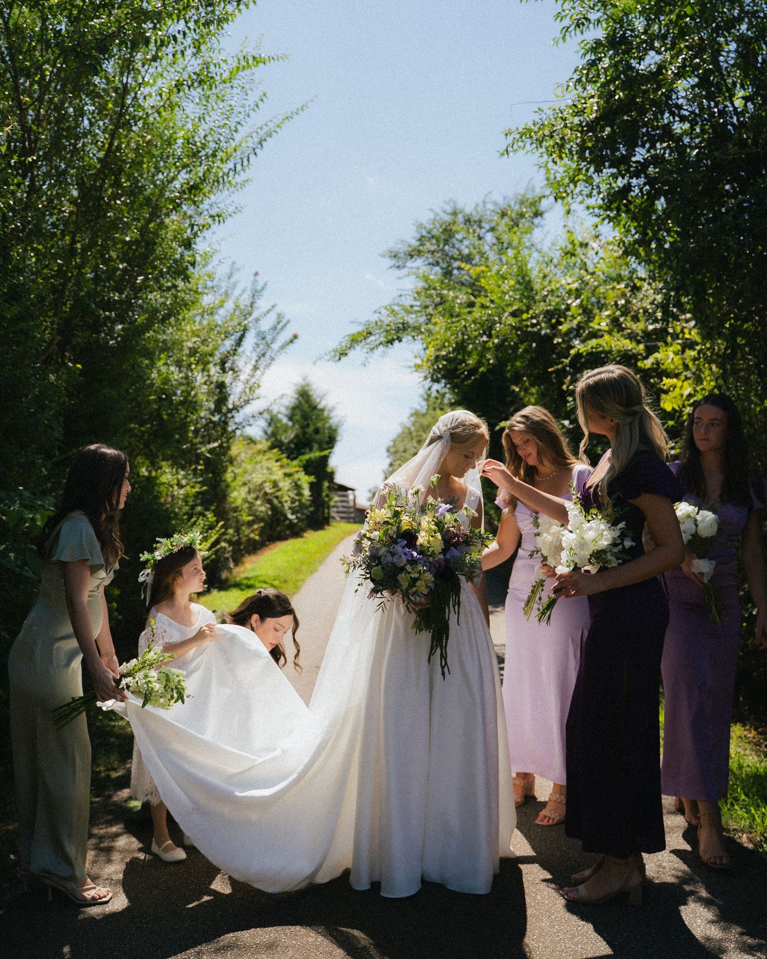 Bride in white gown with veil surrounded by bridesmaids in colorful dresses on sunny tree-lined garden path.