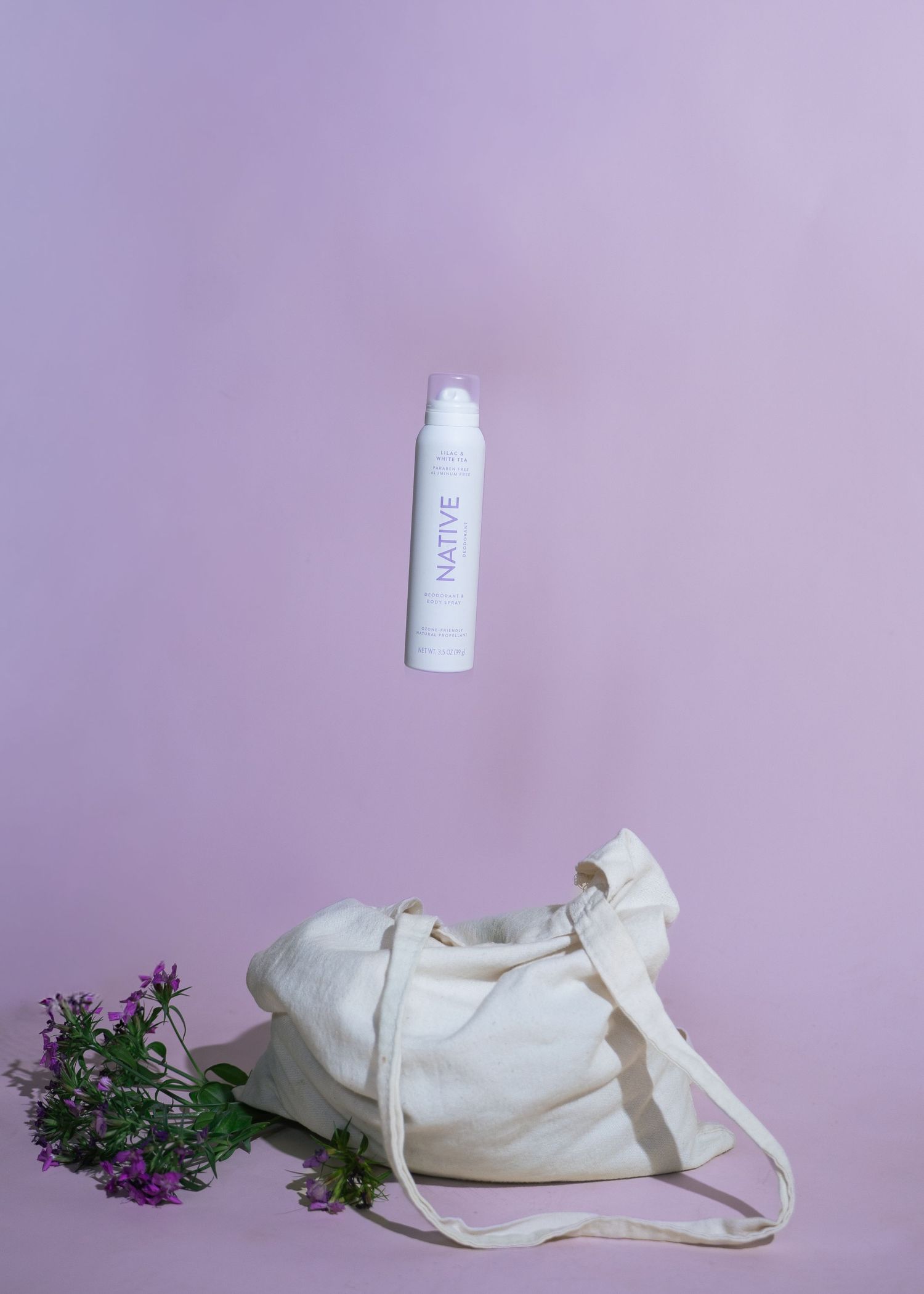 Native deodorant body spray floating above a canvas tote bag with purple flowers on a lavender background.