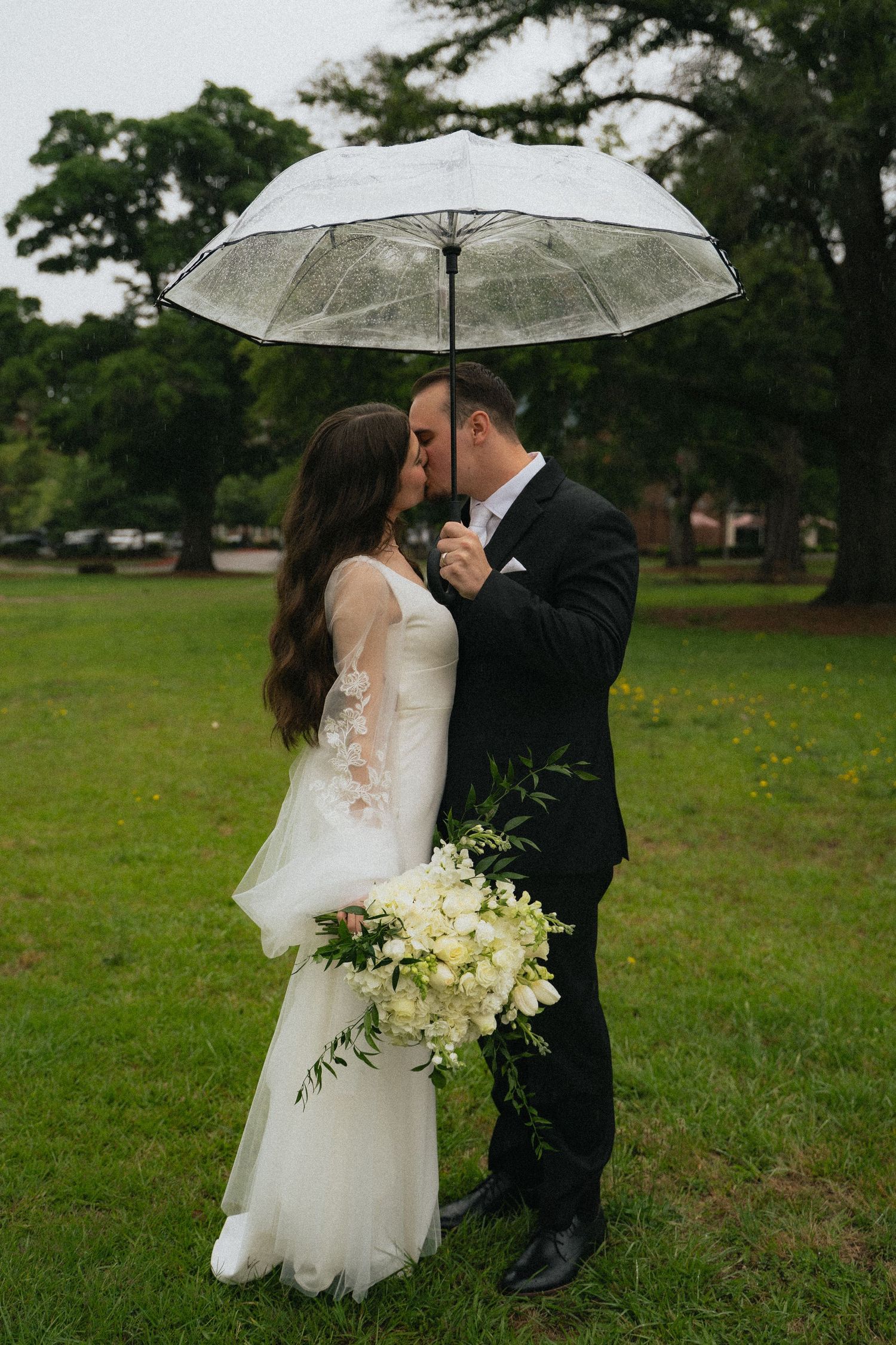 Bride and groom kissing under clear umbrella in the rain, holding white floral bouquet.