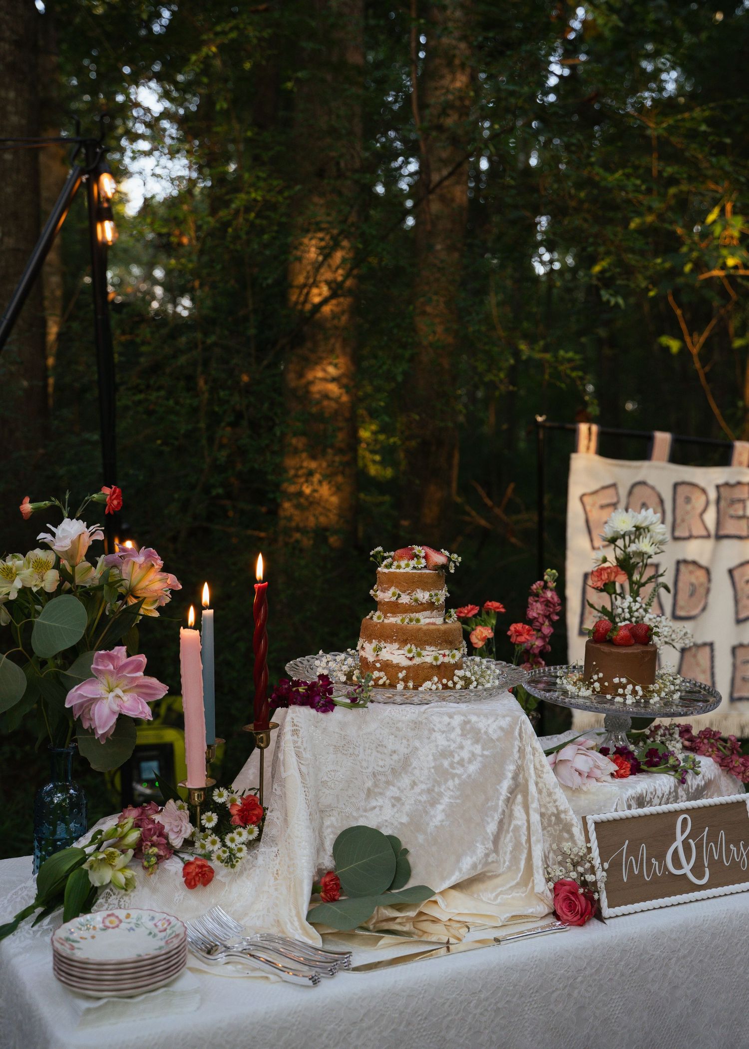 Romantic woodland wedding cake table with lit candles, fresh flowers, and Mr. & Mrs. sign.