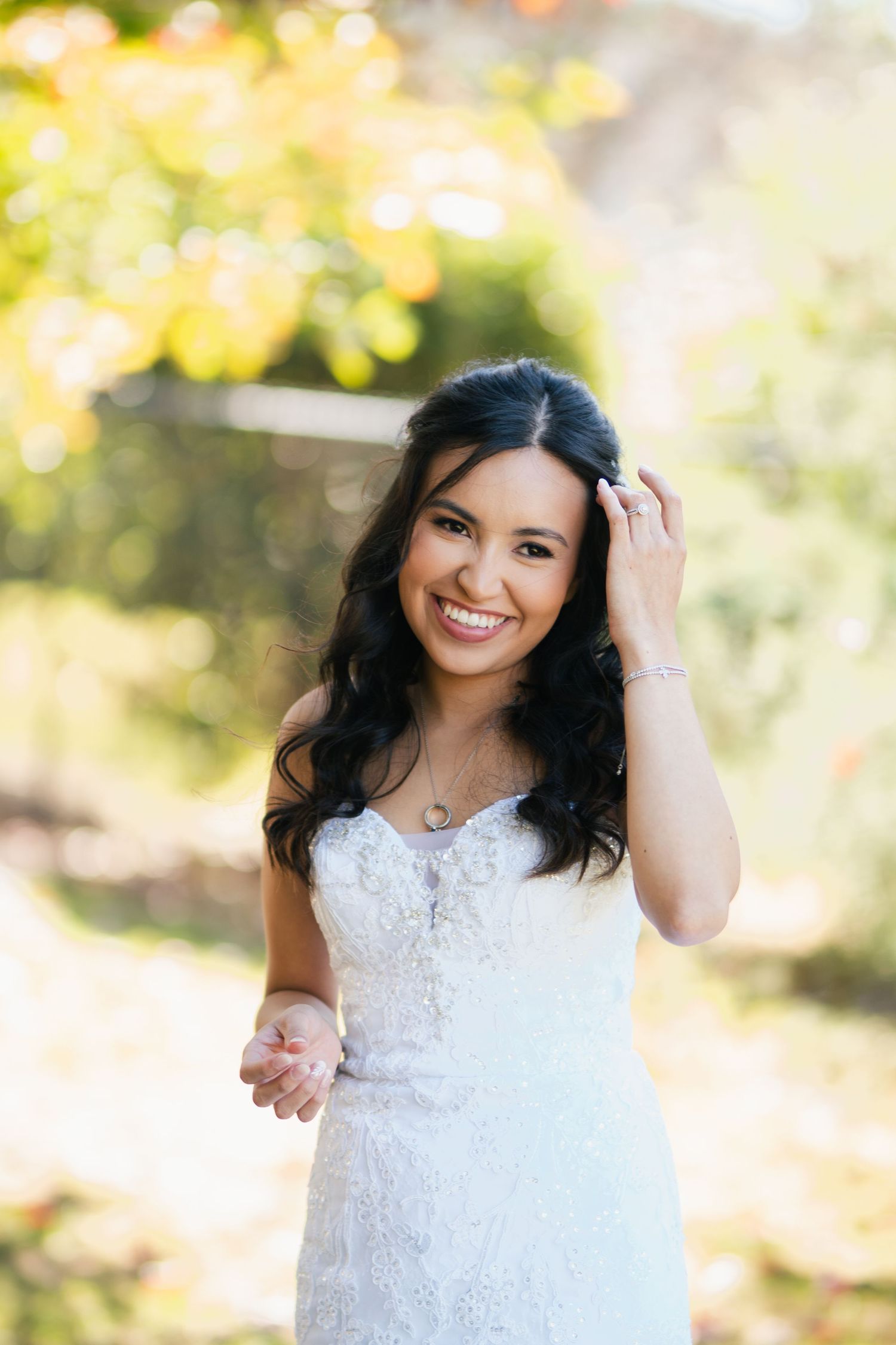 Smiling bride in white lace dress with dark hair poses outdoors in golden autumn light.
