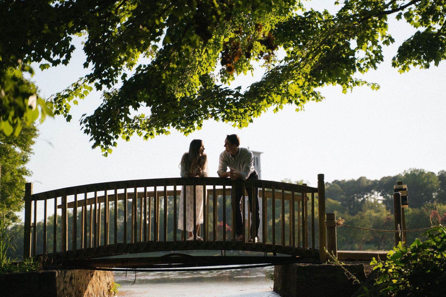 Silhouette of a couple standing on a wooden bridge under a large tree at golden hour.