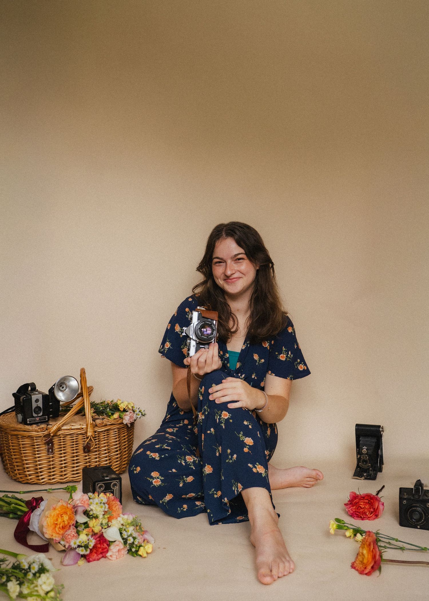Young woman in floral dress sitting with vintage cameras, flowers, and a wicker basket on beige background.