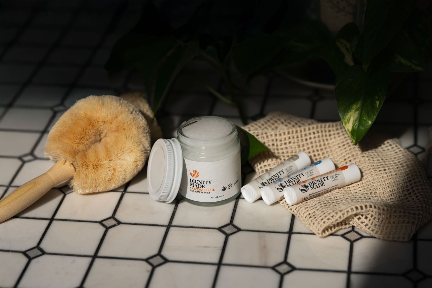 Natural beauty products with cream jar, lip balms, and scrub brush on tile with plant.