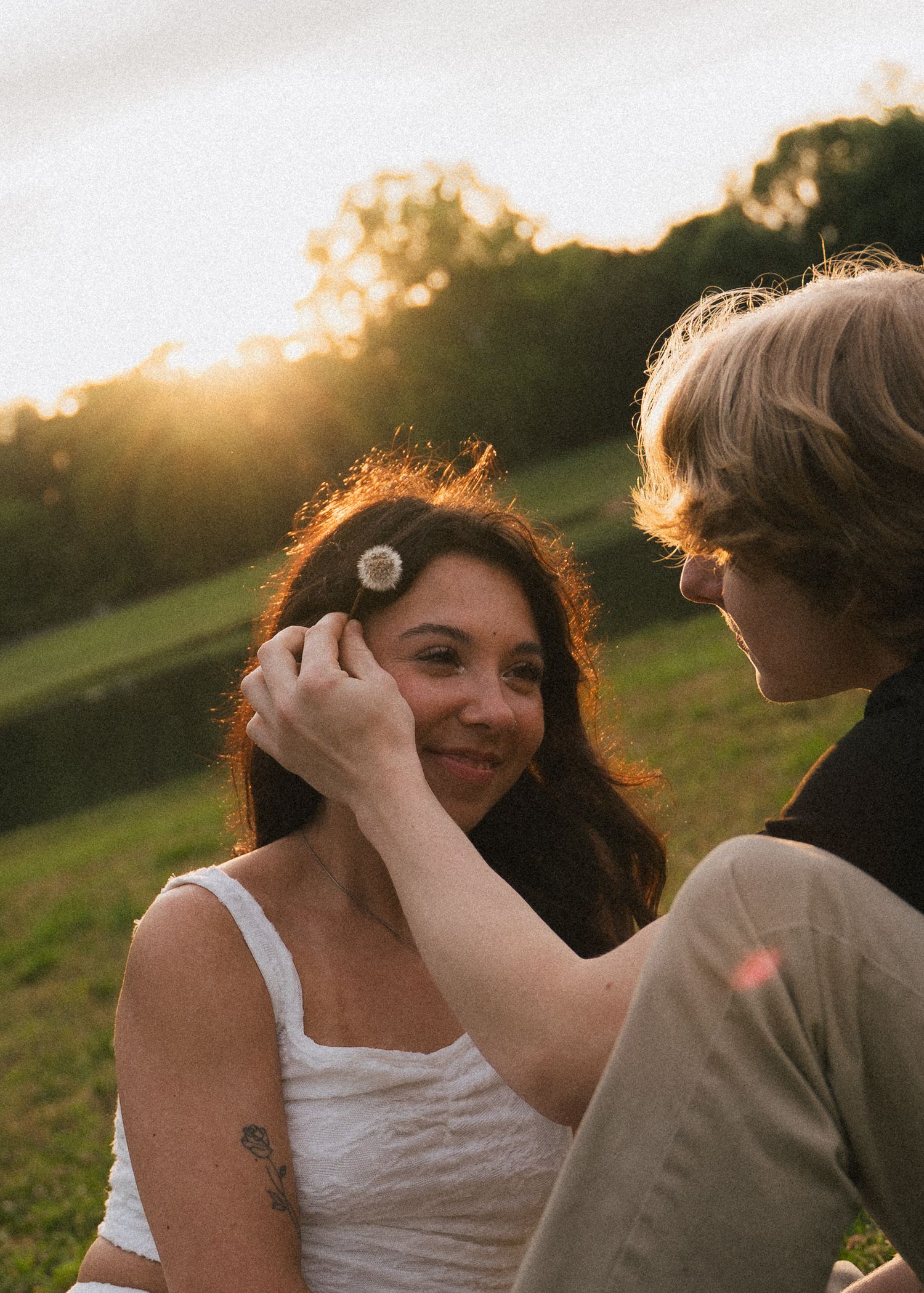 couple sits in a grassy field smiling at each other while the man tucks a old dandelion behind the woman's ear