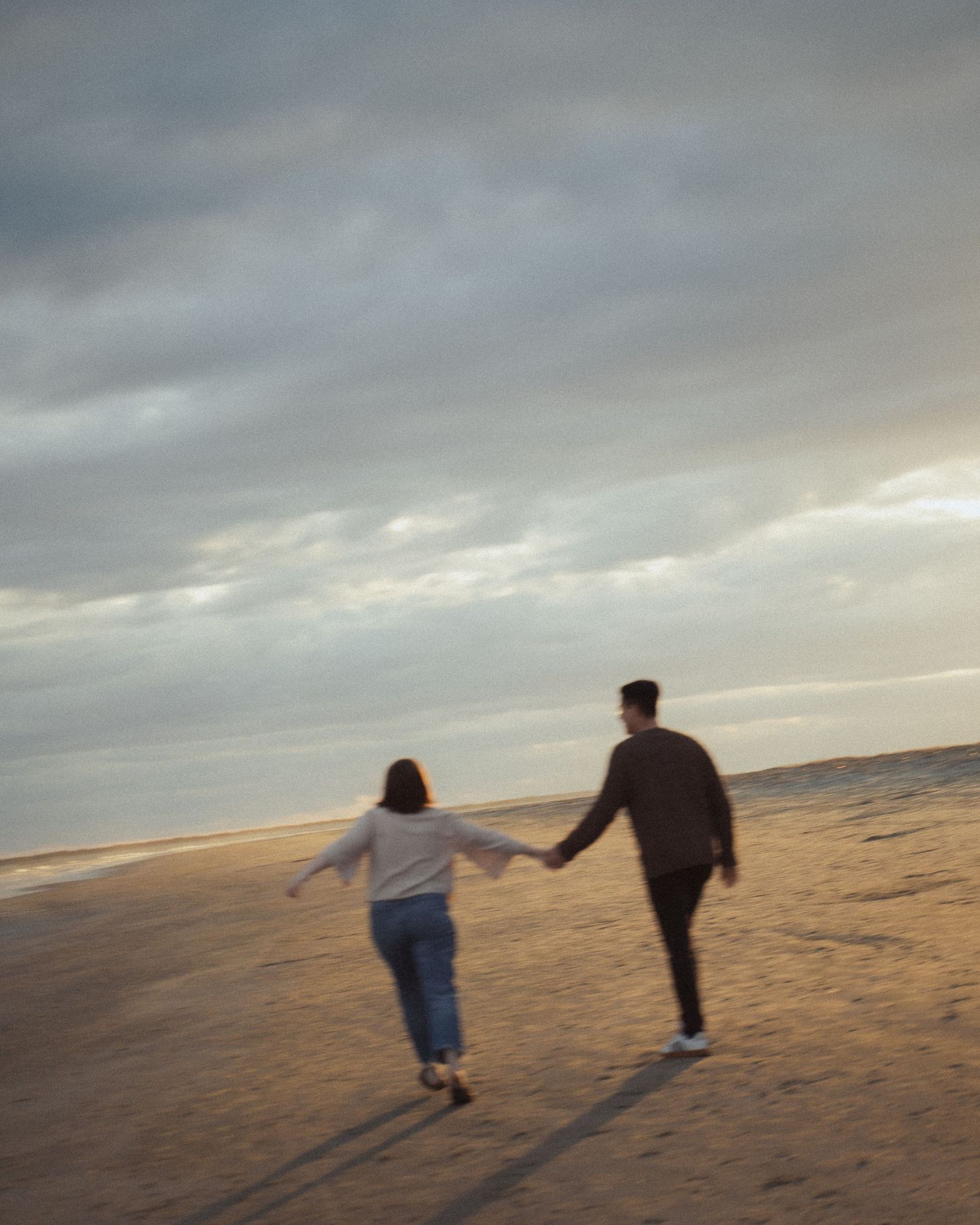 Couple walks hand in hand along a vast sandy beach under a dramatic cloudy sky.