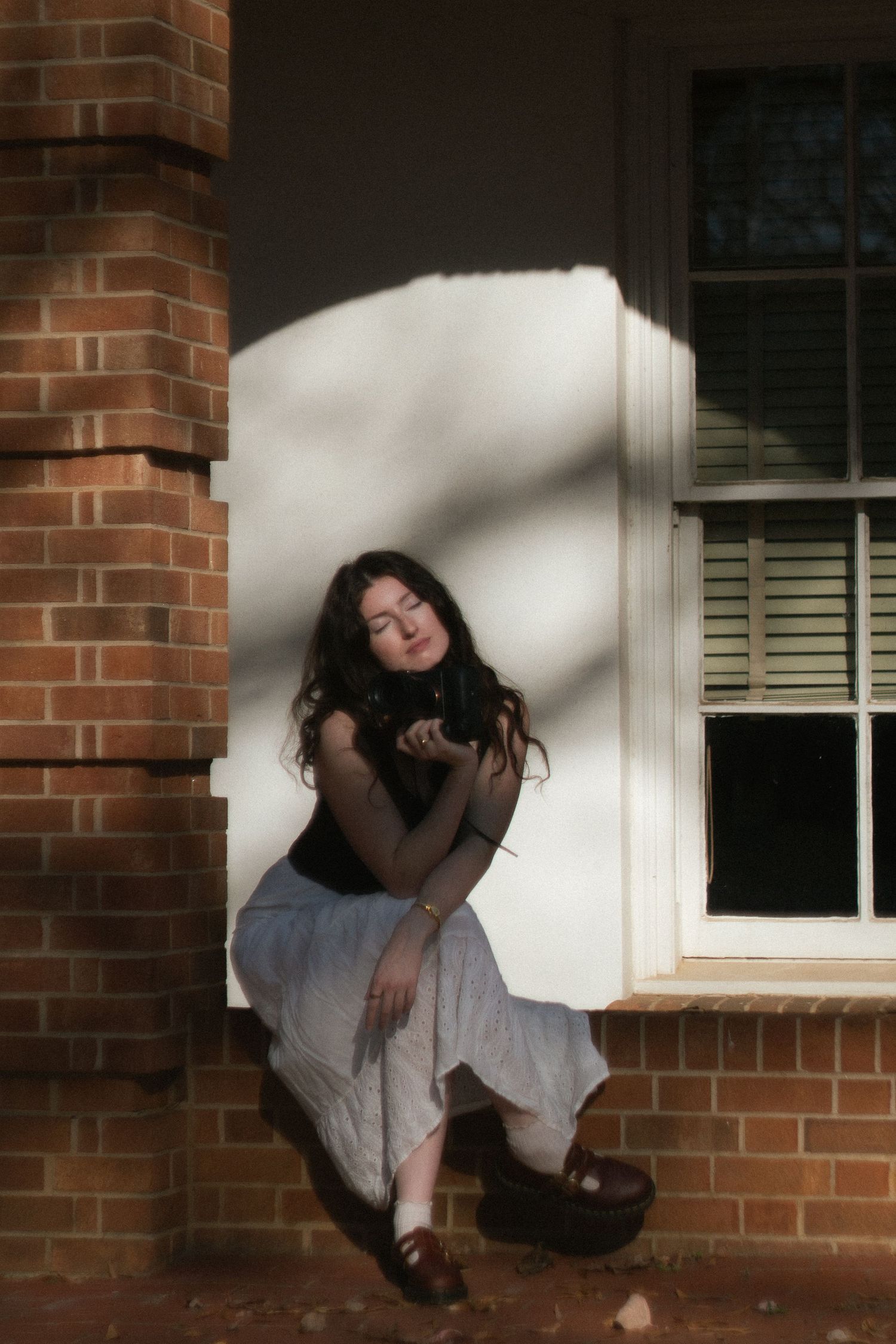 Young woman with dark curly hair sitting by a brick building column in dappled sunlight, eyes closed.