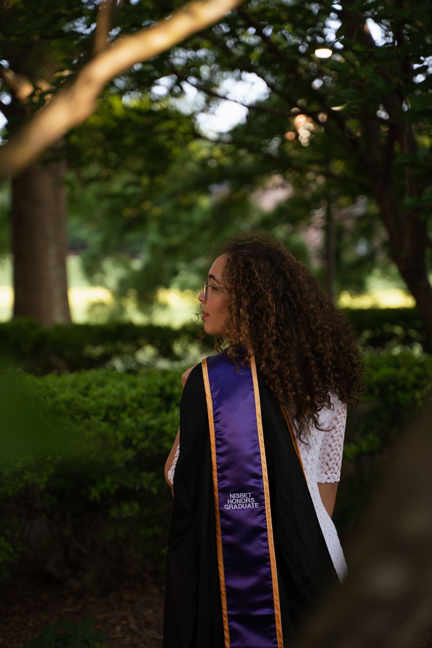 Young woman in graduation stole gazing thoughtfully among lush green trees and hedges.