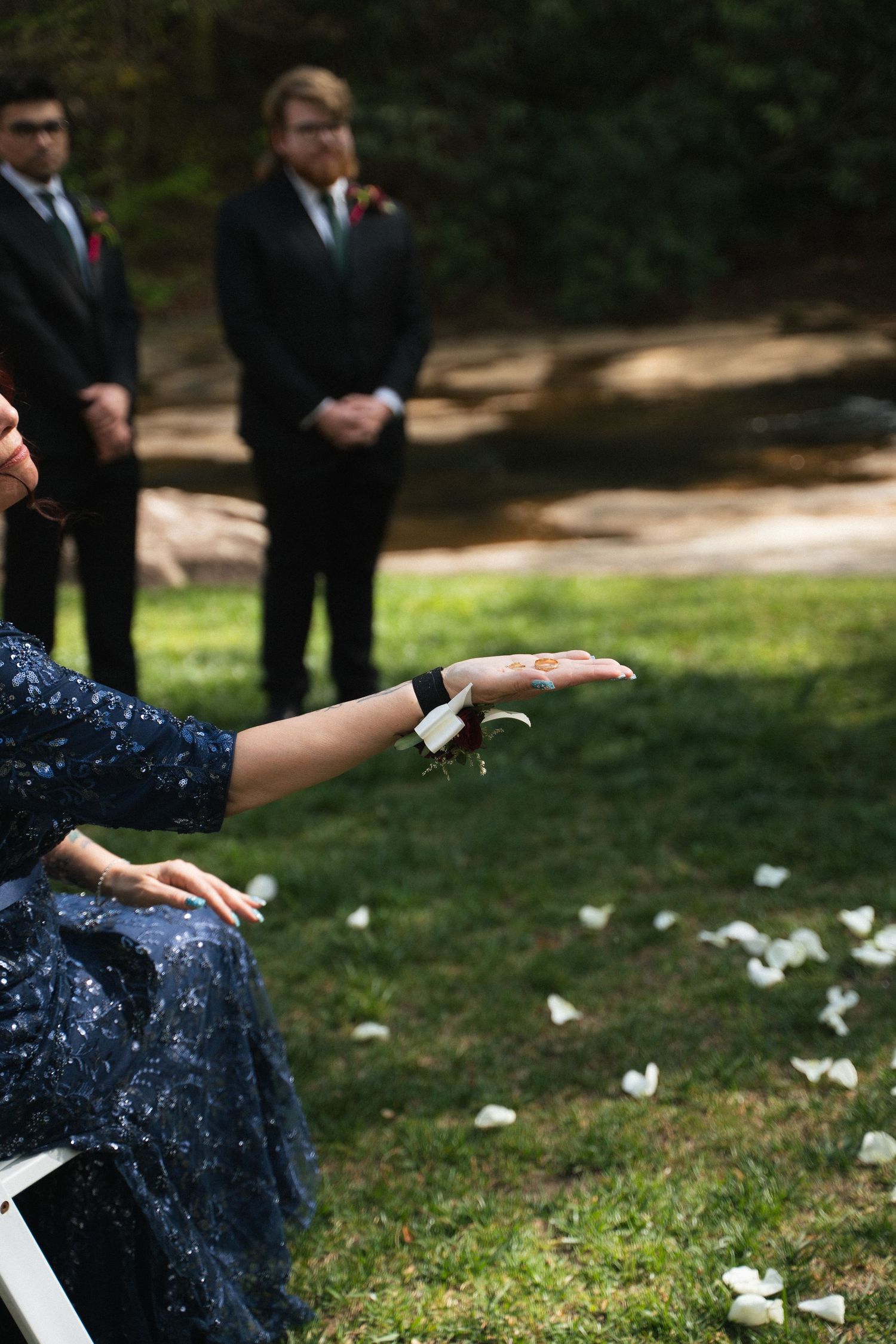 Flower girl tosses white petals at outdoor garden wedding ceremony with groomsmen standing nearby.