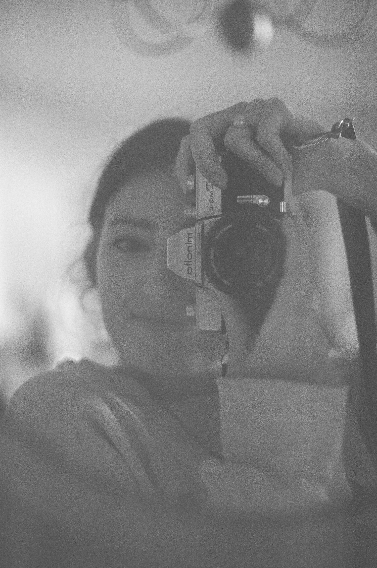 Black and white self-portrait of a woman holding a vintage film camera up to her face in a mirror reflection.