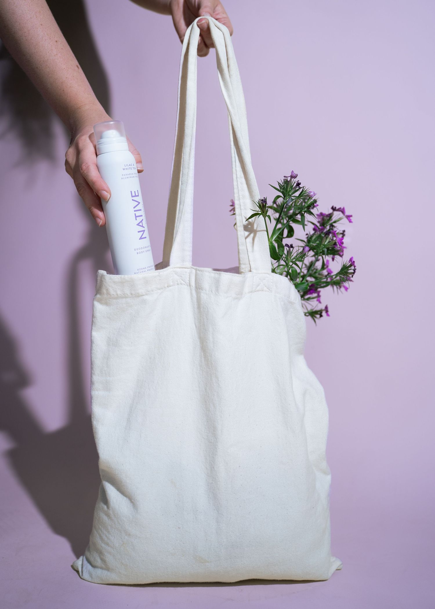 Hands holding canvas tote bag with Native deodorant spray and fresh purple wildflowers on pink background.
