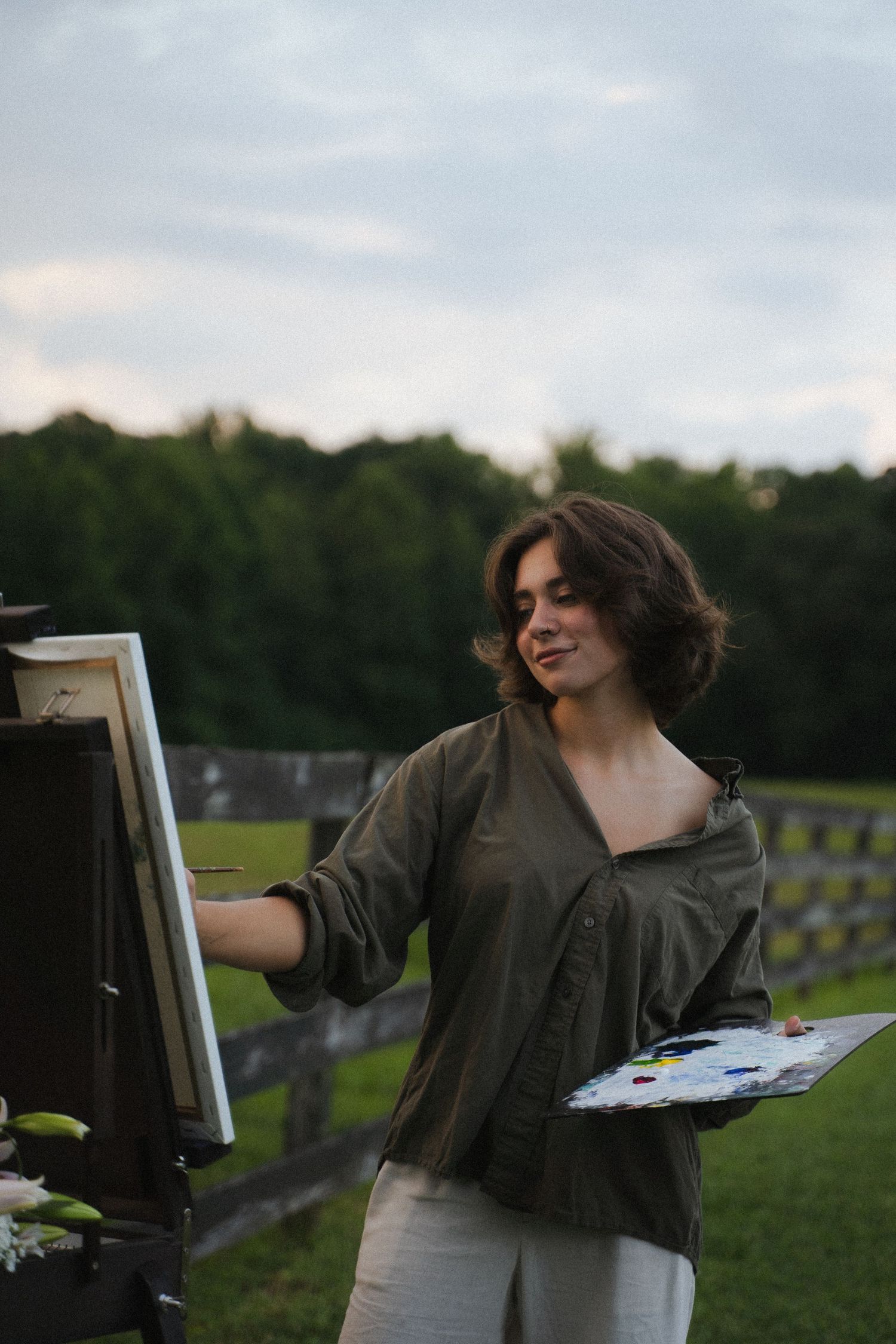 Young woman in olive jacket painting on an easel outdoors at dusk near a wooden fence.