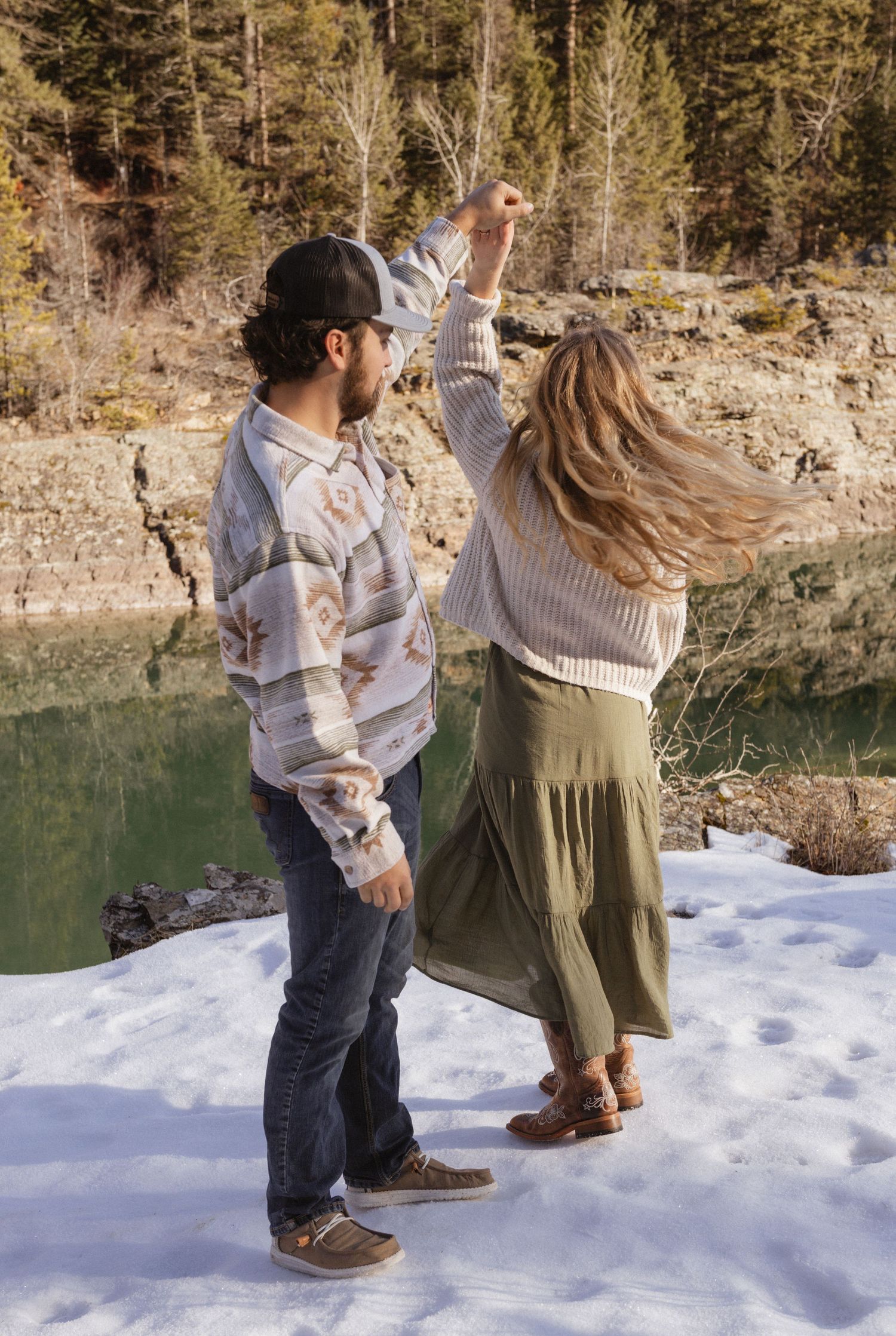 An engaged couple twirling around in the snow next to a river in West Glacier Montana