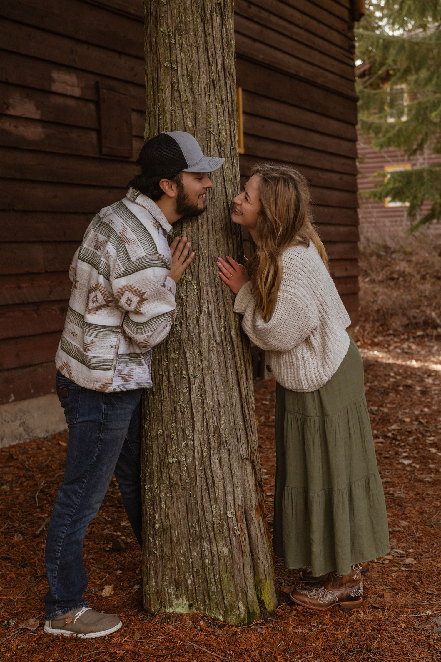 A couple looking at each other around a tree during an engagement session at lake McDonald in West Glacier Montana